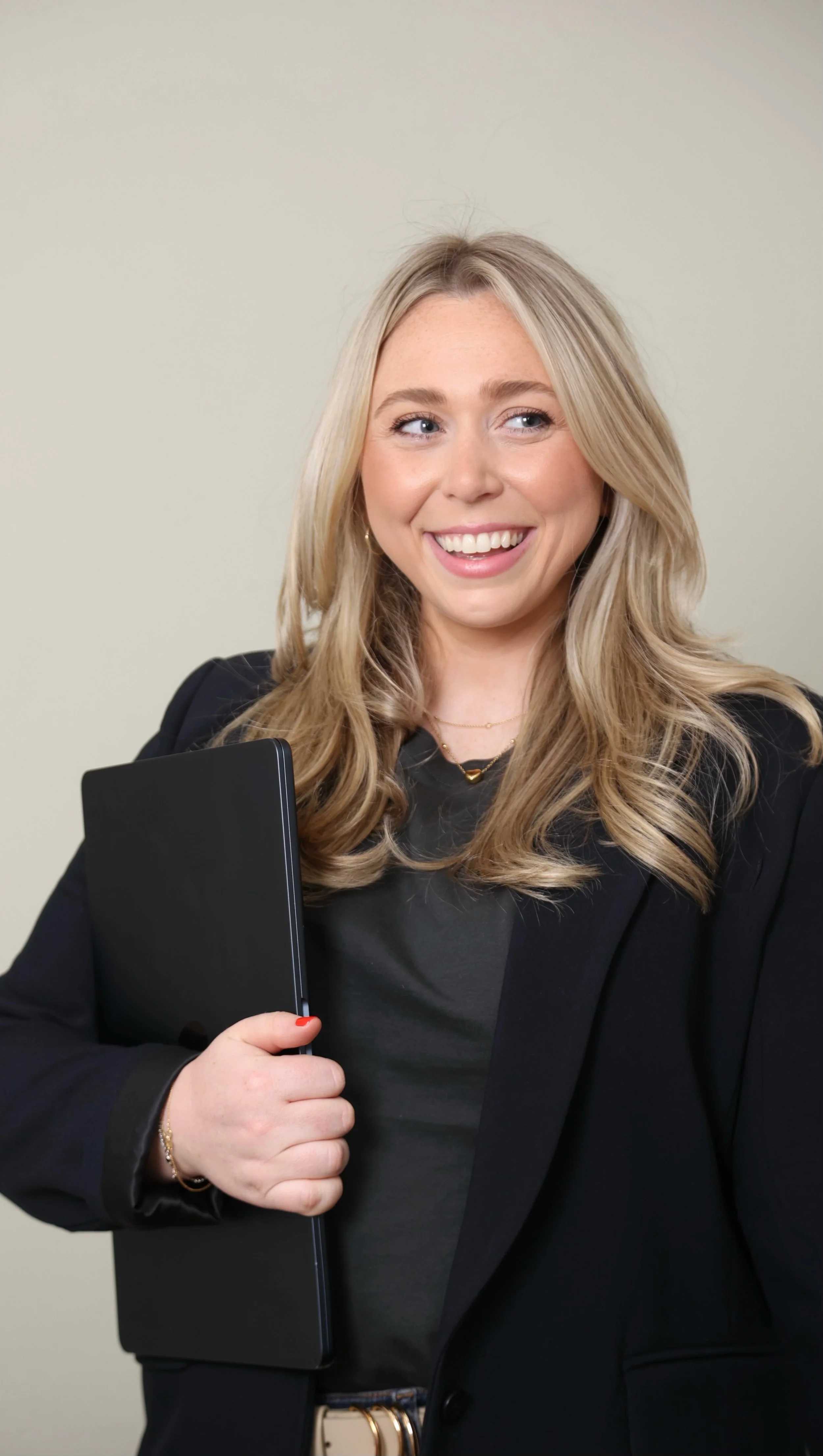 A young woman with blonde hair, a dark blazer, and a necklace, smiling and holding a closed black notebook in her left hand.