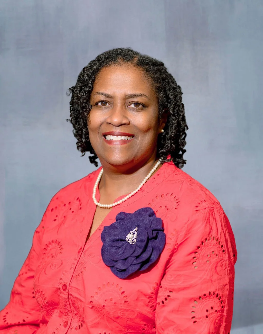 A portrait of an African American woman smiling, wearing a red dress with detailed embroidery, a pearl necklace, and a large navy blue fabric flower with a brooch on her left chest, set against a plain grey background.