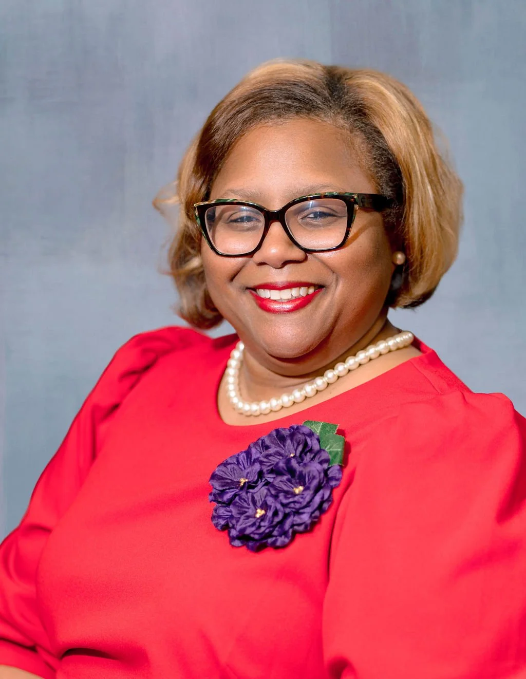 A woman with glasses and pearl jewelry smiling in a red dress with a purple flower accessory on her chest.