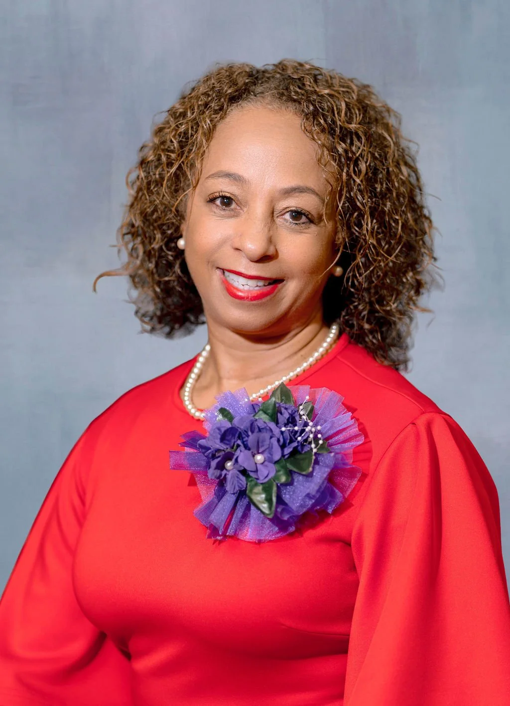 A woman with curly hair wearing a red dress, a pearl necklace, and pearl earrings, holding a purple flower corsage, smiling at the camera, with a plain gray background.