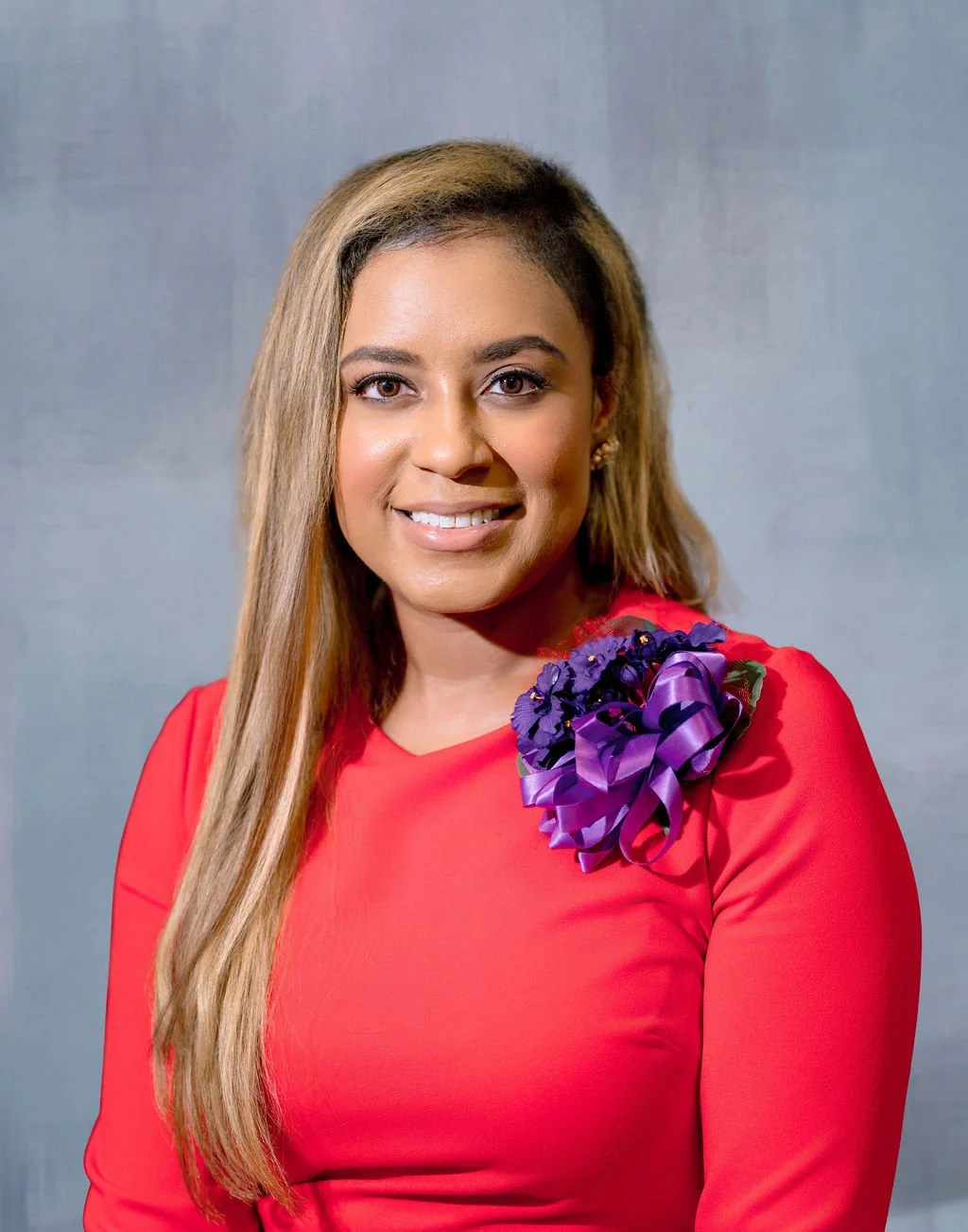A woman with long, styled hair, wearing a red dress with a purple floral embellishment on her shoulder, smiling against a plain grey background.