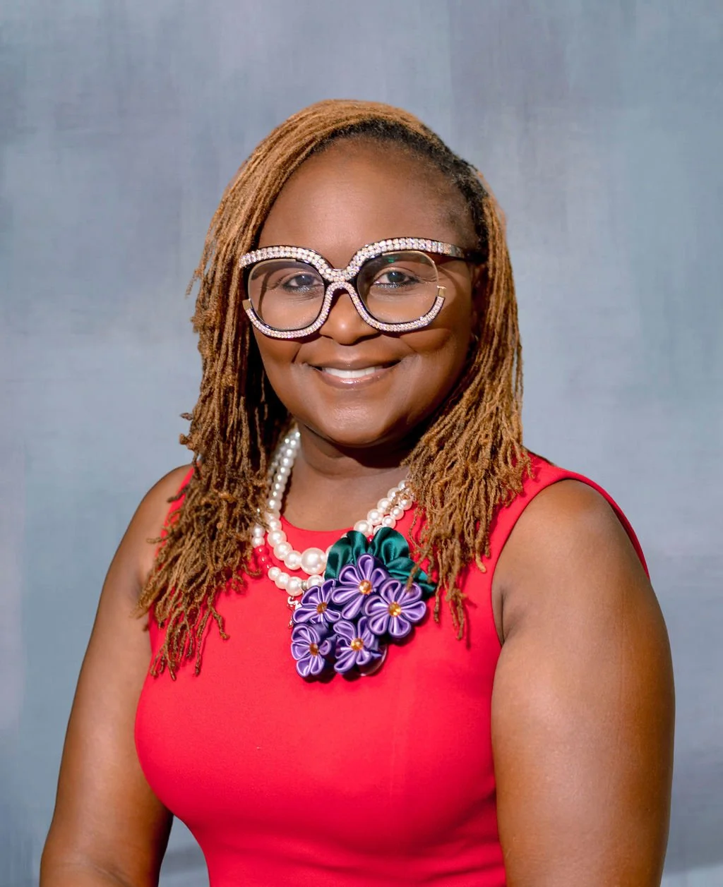 A woman with brown dreadlocks smiling, wearing large rhinestone eyeglasses, a red sleeveless top, double strand pearl necklace, and a colorful floral statement necklace against a light gray background.