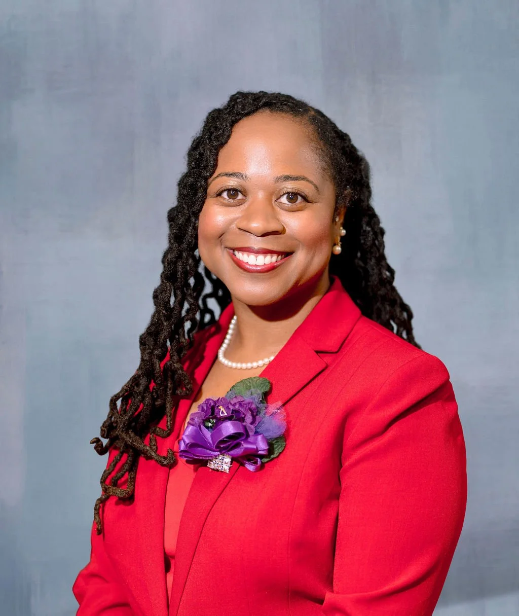 A smiling woman with dark, twisted dreadlocks wearing a red blazer, pearl necklace, pearl earrings, and a purple boutonniere, against a plain gray background.