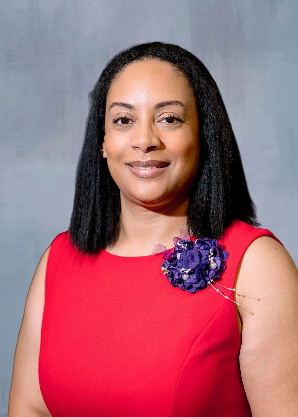 A woman with shoulder-length black hair wearing a sleeveless red dress with a purple floral brooch on her left shoulder, smiling against a plain gray background.