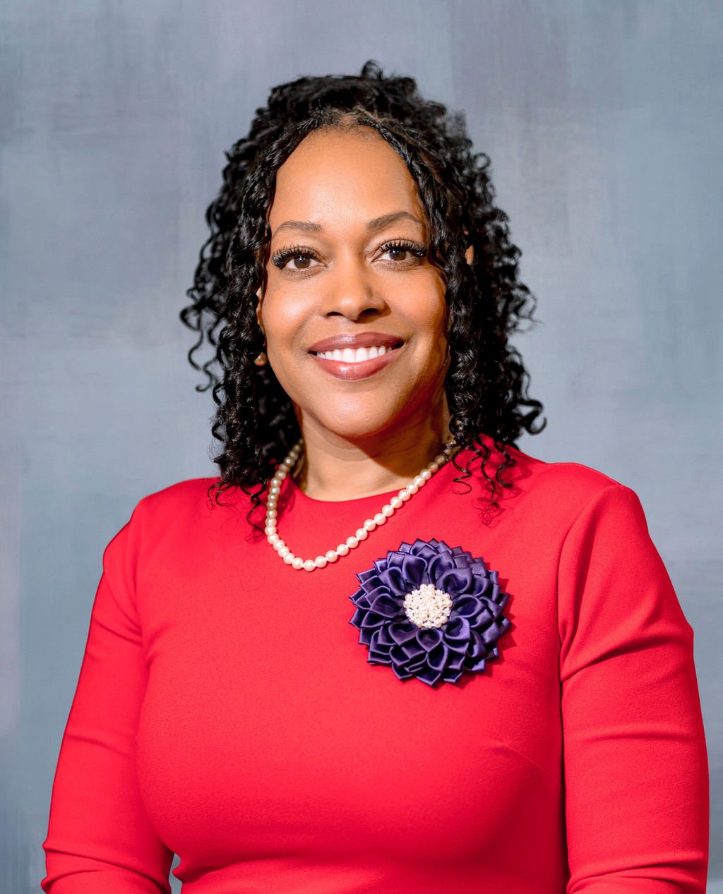 A woman with curly black hair smiling, wearing a red dress, a pearl necklace, and a large dark purple fabric flower brooch on her left chest, posed against a gray background.