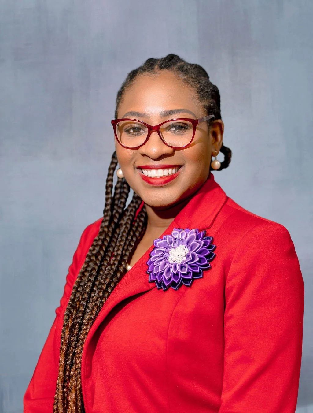 A woman with braided hair, wearing red glasses, red blazer, pearl earrings, a purple flower pin, and red lipstick, smiling at the camera against a gray background.