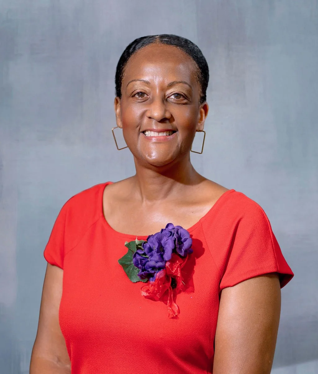 A woman with short black hair, wearing a red dress with a purple flower and green leaf accessory pinned to her chest, smiling against a plain background.