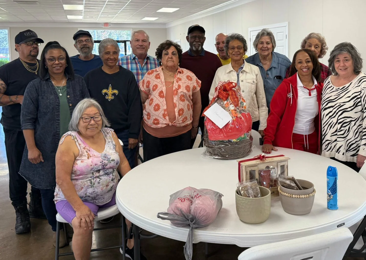 Group of diverse elderly people gathered indoors, smiling for a photo, with a round table in front of them holding a gift basket, wrapped presents, and various items, in a community or event setting.