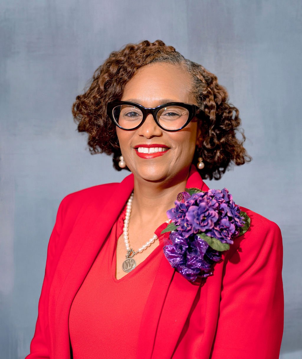 A portrait of a woman with curly brown hair wearing glasses, a red blazer with a purple corsage, pearl earrings, and a pearl necklace, smiling against a neutral background.