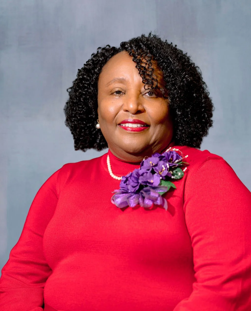 A woman with short curly black hair wearing a red dress and a pearl necklace, with purple flowers attached to her dress, smiling at the camera against a plain gray background.