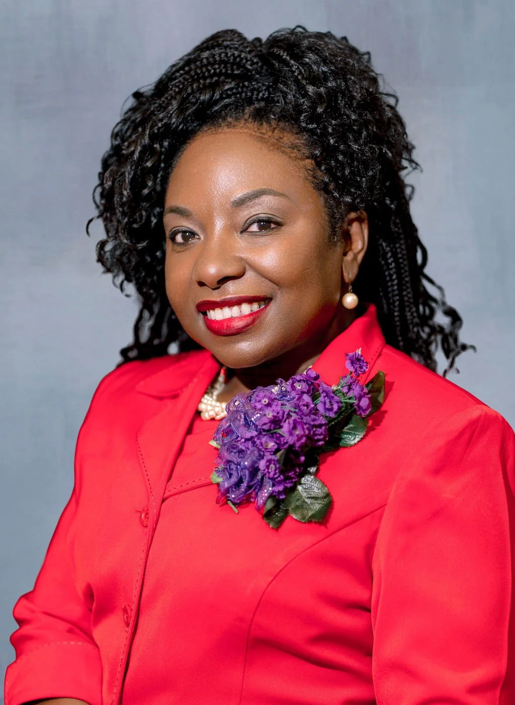 A woman with dark, curly hair and dark skin, wearing a red outfit with a purple floral brooch and pearl jewelry, smiling at the camera against a neutral background.