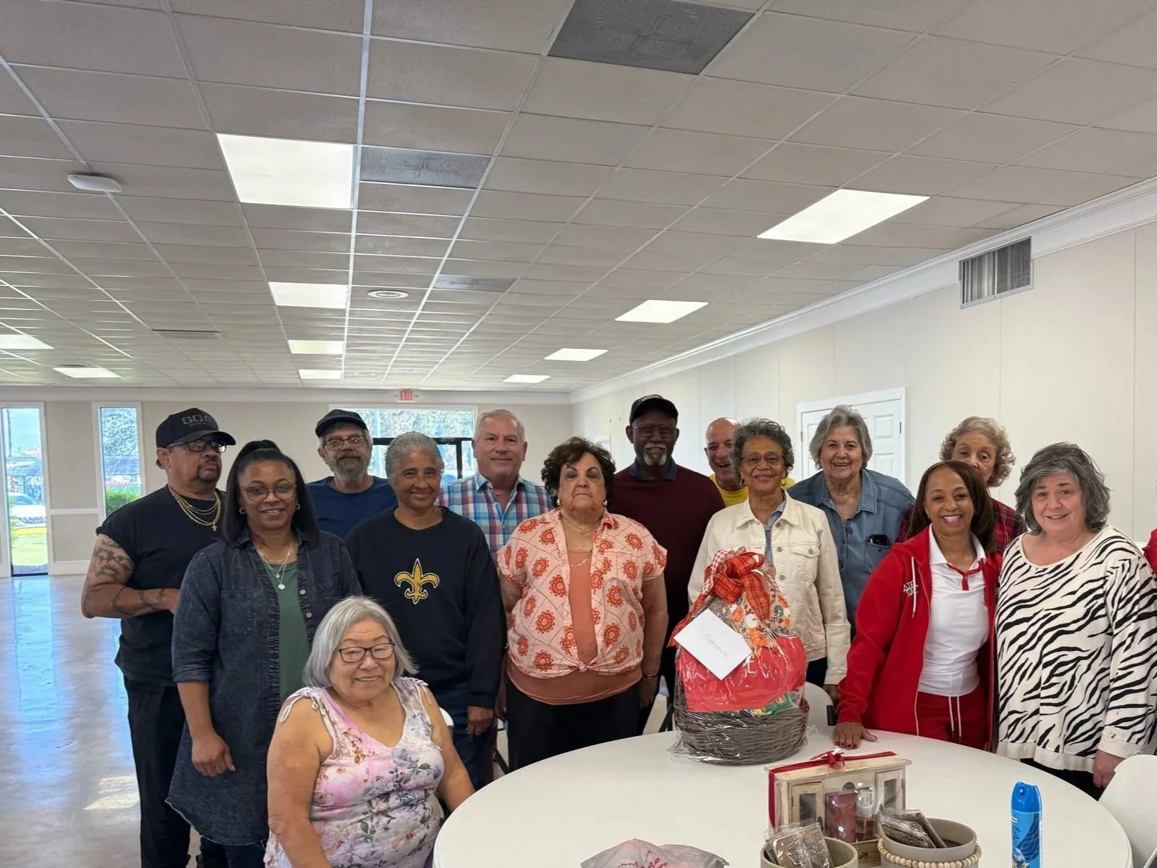 A group of diverse elderly people gathered around a white table in a community room, celebrating with a red and white gift basket and smiling at the camera.
