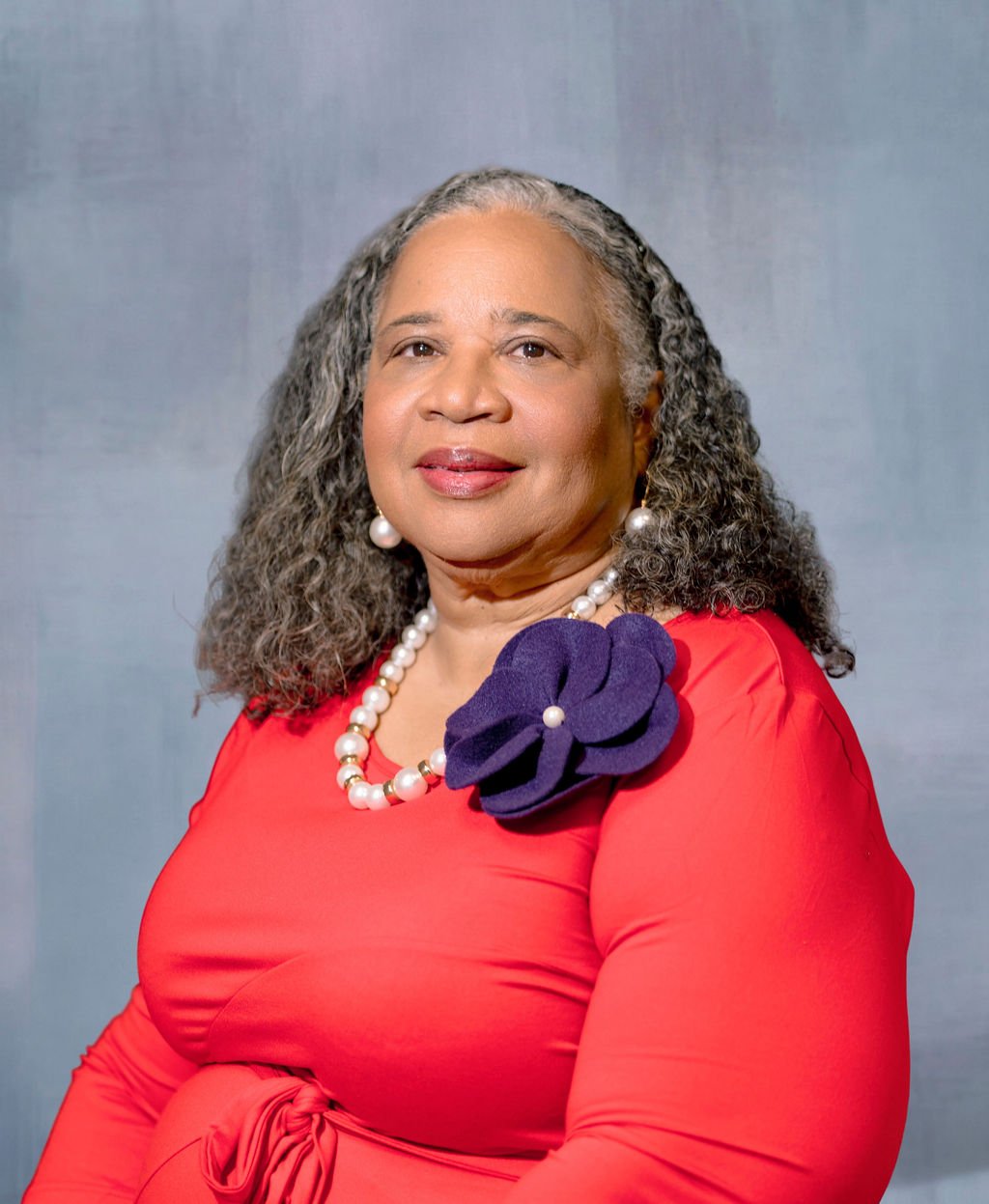 A portrait of an older woman with curly gray hair, wearing a red dress with a purple fabric flower on her shoulder, pearl earrings, and a pearl necklace, standing against a plain gray background.