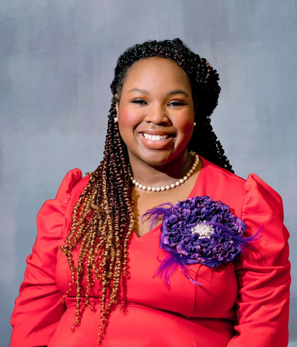 A smiling Black woman with long braided hair, wearing a red dress with puffed sleeves, a pearl necklace, pearl earrings, and a large purple floral brooch on her chest.