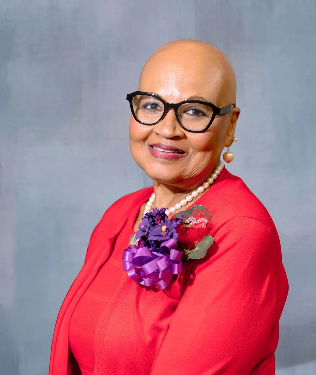 A woman with a bald head, wearing black glasses, a red outfit, a pearl necklace, and earrings. She has a purple flower corsage on her red outfit and is smiling at the camera against a neutral background.