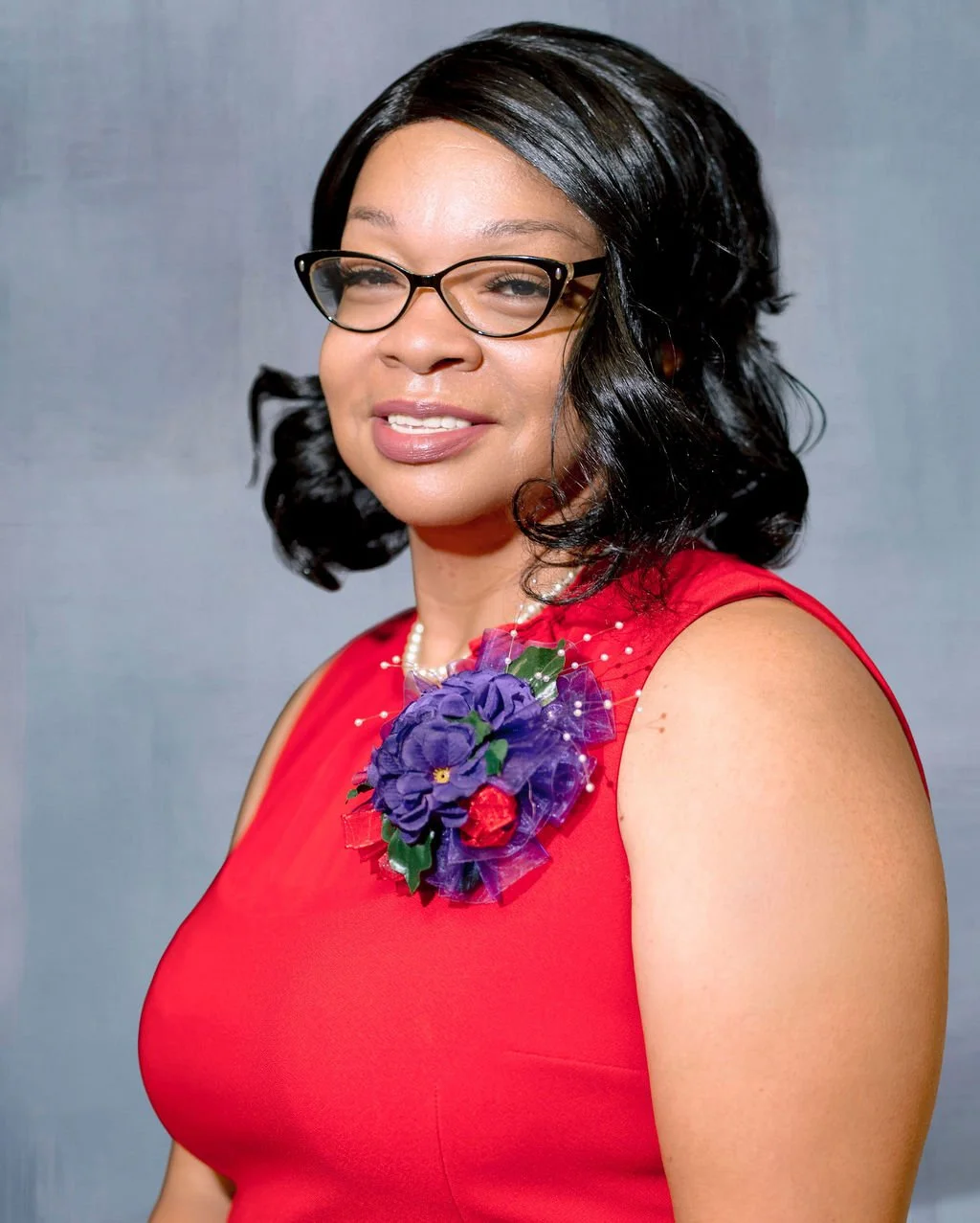 A woman with black hair, glasses, wearing a red dress with a purple and red floral brooch on her shoulder, smiling against a plain background.