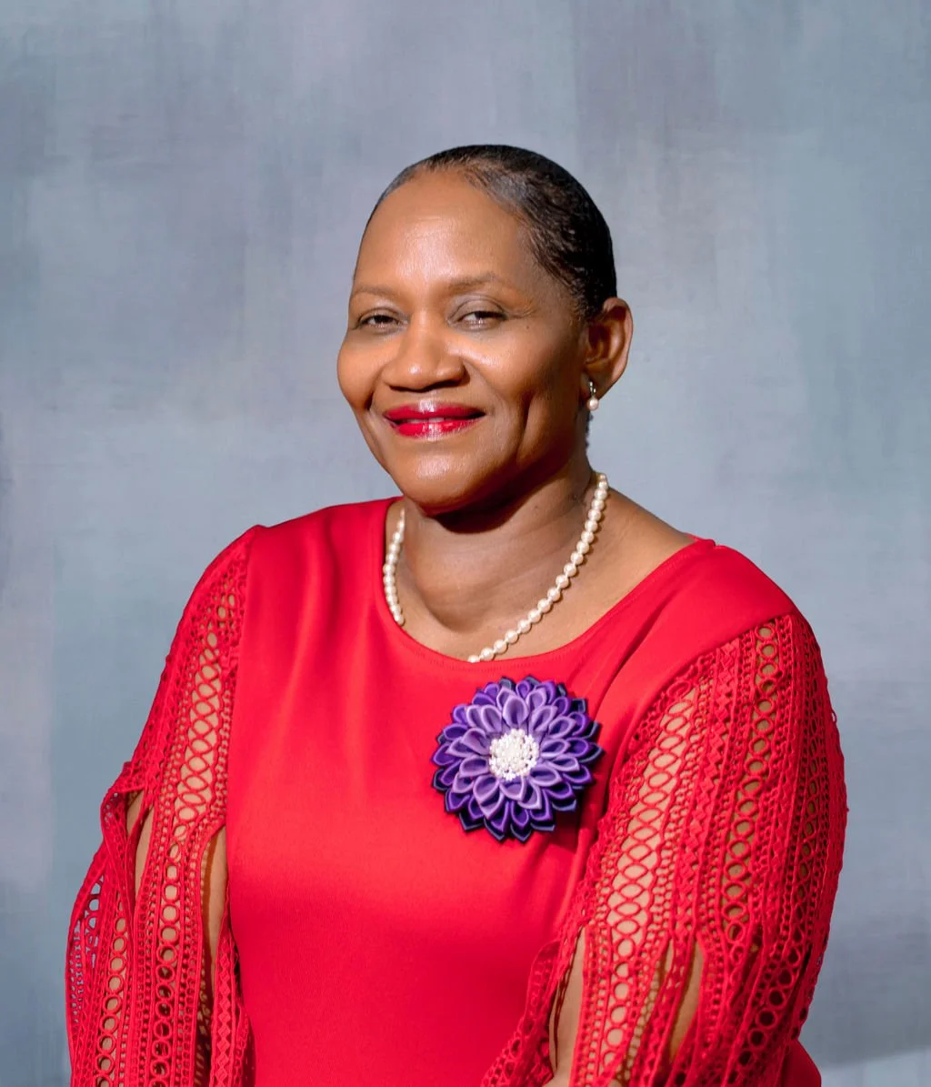 A smiling African American woman with short hair, wearing a red dress with lace details on the sleeves, a pearl necklace, pearl earrings, and a large purple floral brooch.