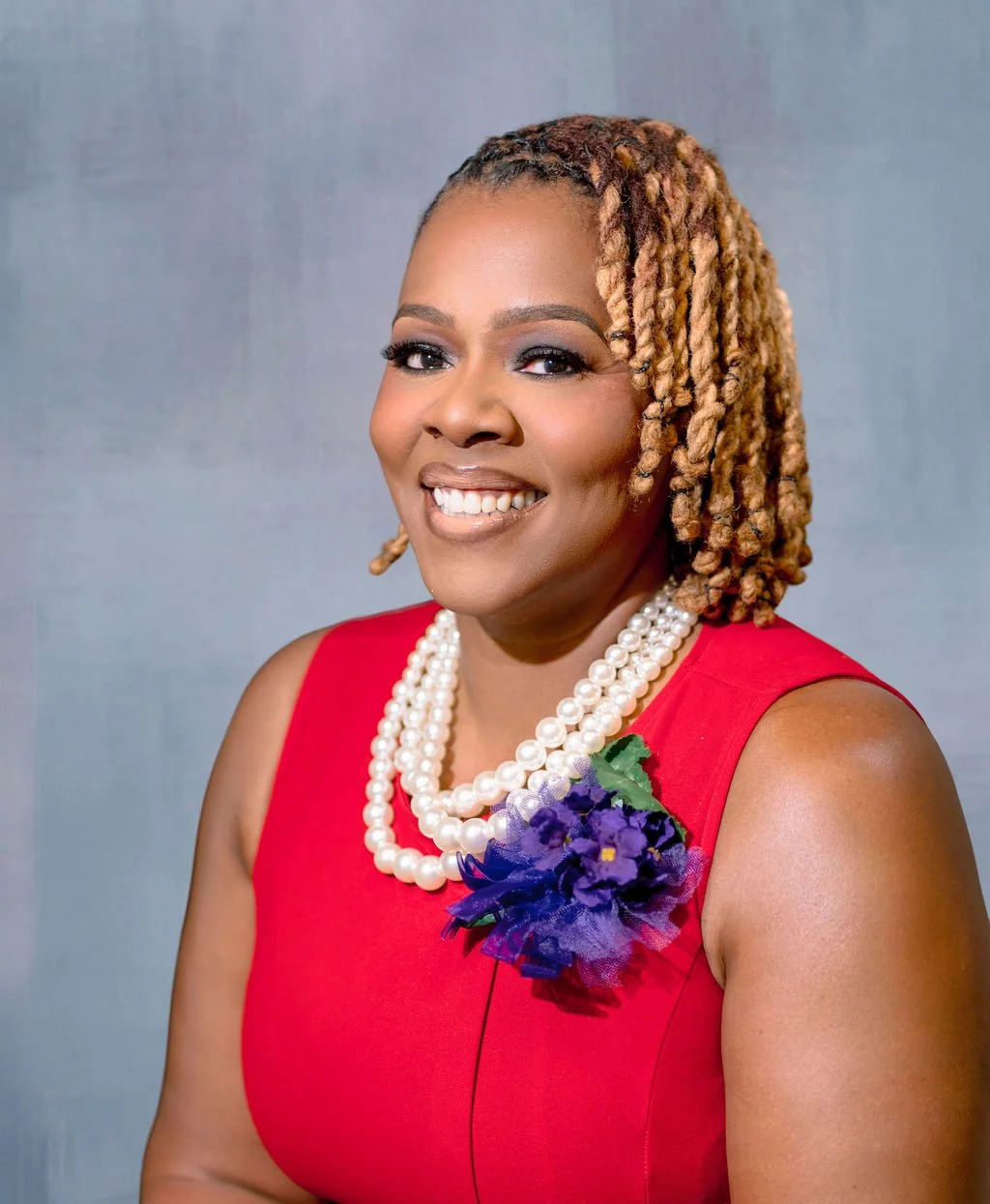 A smiling woman with dreadlocks wearing a red dress, a pearl necklace, and a purple floral brooch.