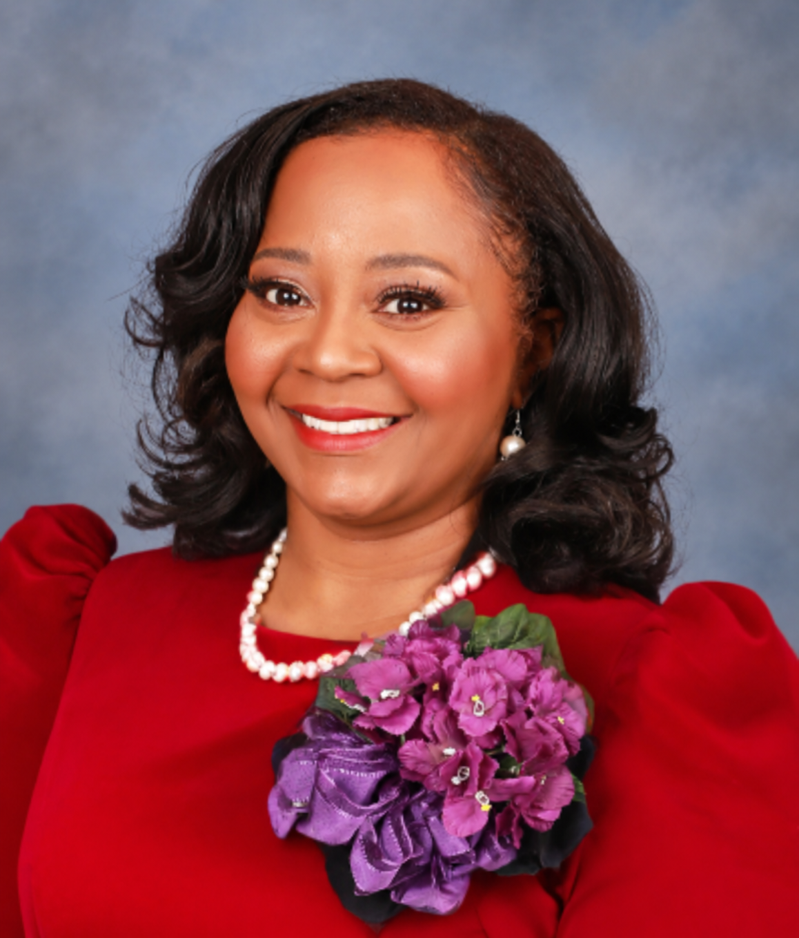 A woman with dark wavy hair smiling, wearing a red dress with puffed sleeves, pearl earrings and necklace, holding a purple flower corsage, against a blue and gray background.
