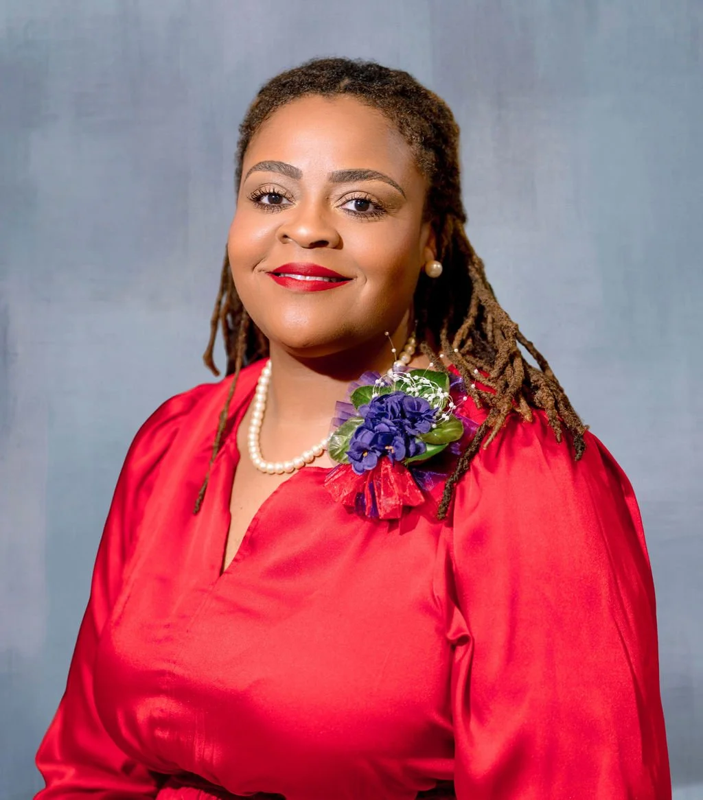 A woman wearing a red dress with a pearl necklace, earrings, and a floral corsage on her shoulder. She has styled dreadlocks and is smiling against a neutral background.