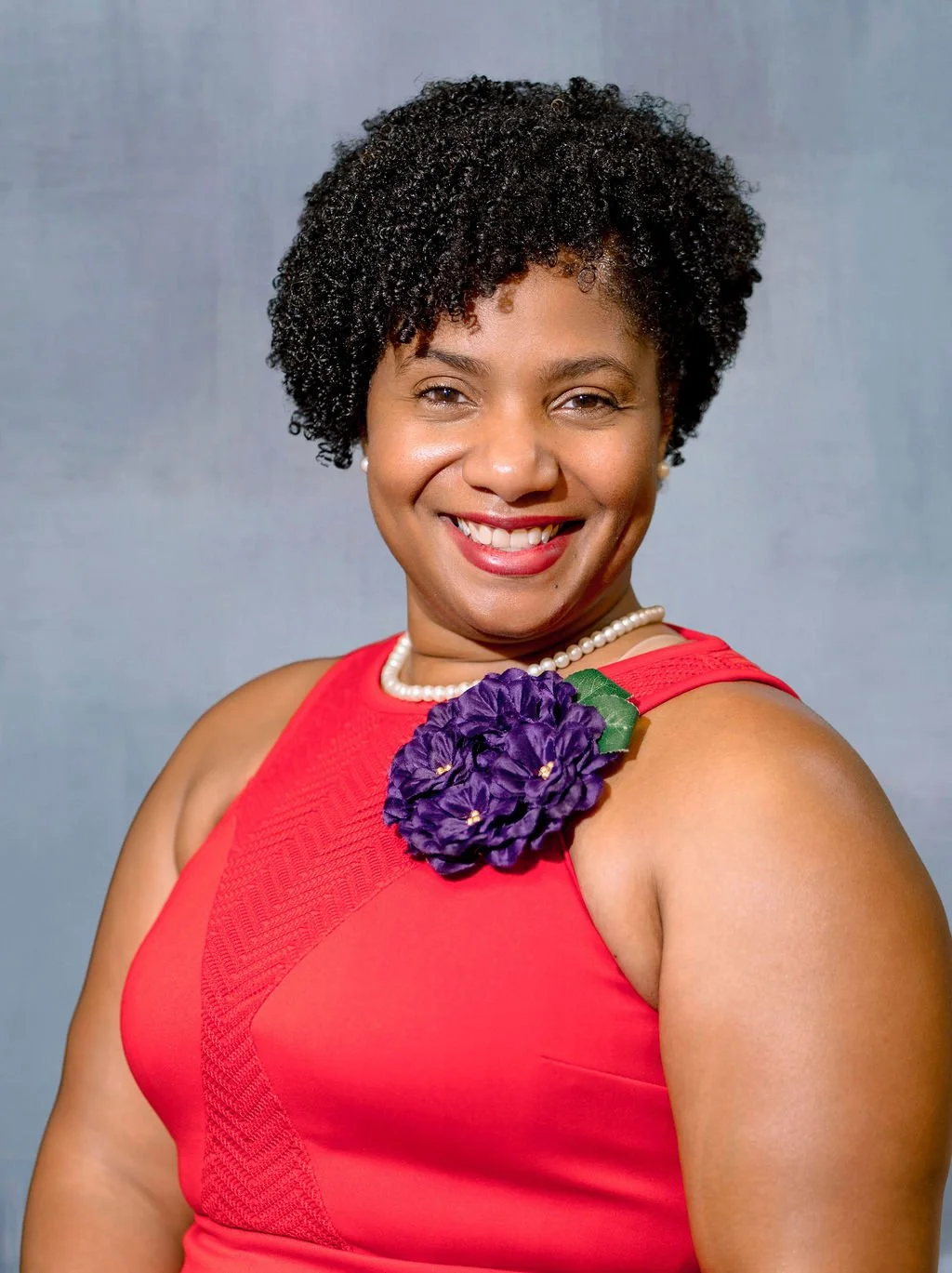 A woman wearing a red dress with a purple flower accessory and a pearl necklace, smiling at the camera against a gray background.