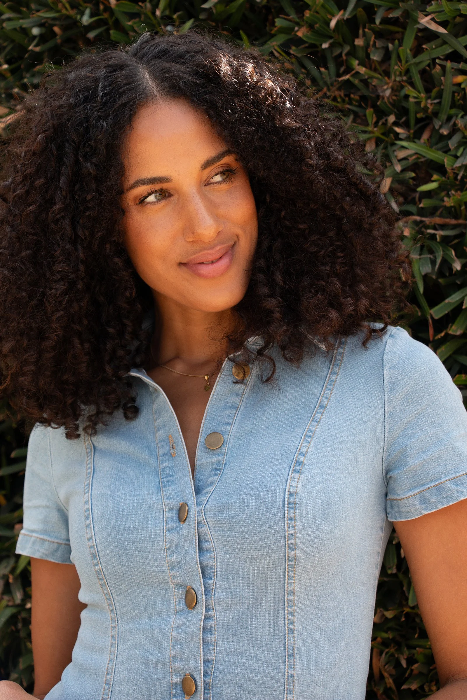 A woman with curly dark hair and light skin wearing a light blue denim shirt, standing outdoors in front of greenery.