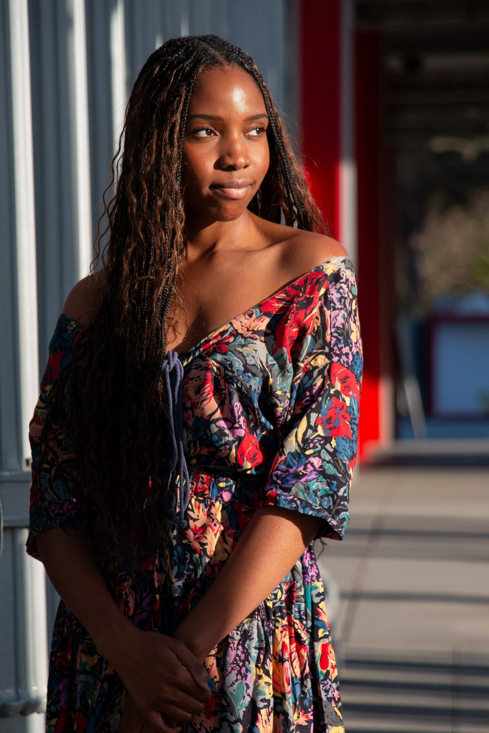 A young woman with long, curly hair wearing a colorful off-shoulder dress with a floral pattern, standing outdoors in front of a metal structure with sunlight illuminating her face.