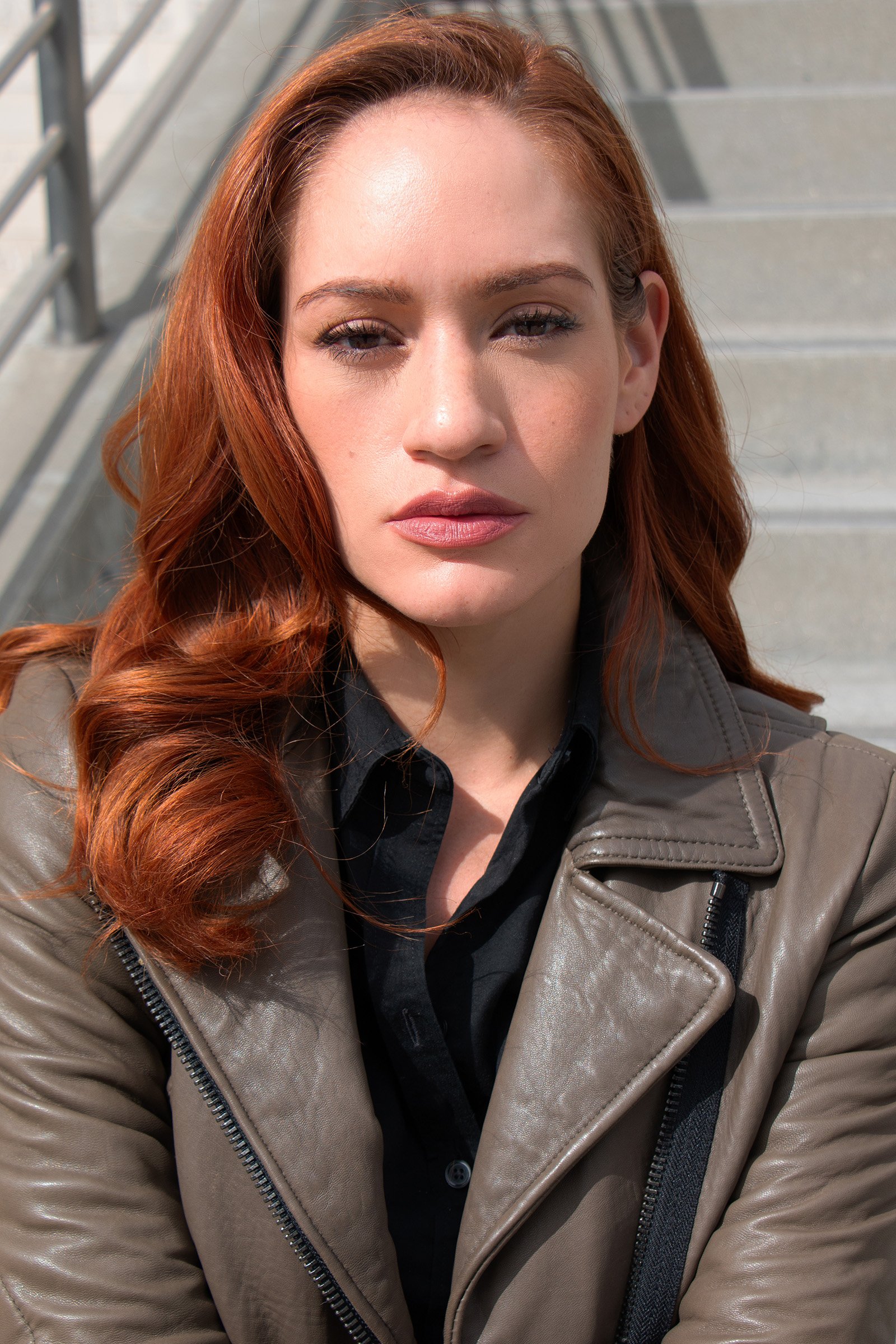 Close-up of a woman with red hair wearing a tan leather jacket and black shirt, standing outdoors on steps.