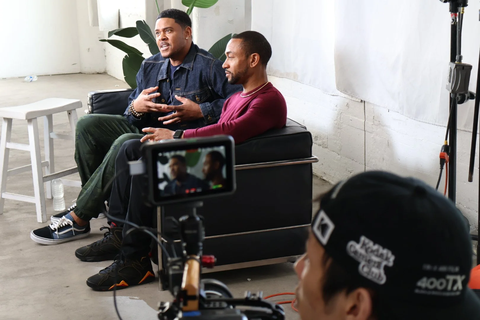 Two men seated on a black couch, engaging in a conversation during a recording session inside a room with white walls and green plants. A camera and crew are visible capturing the scene.