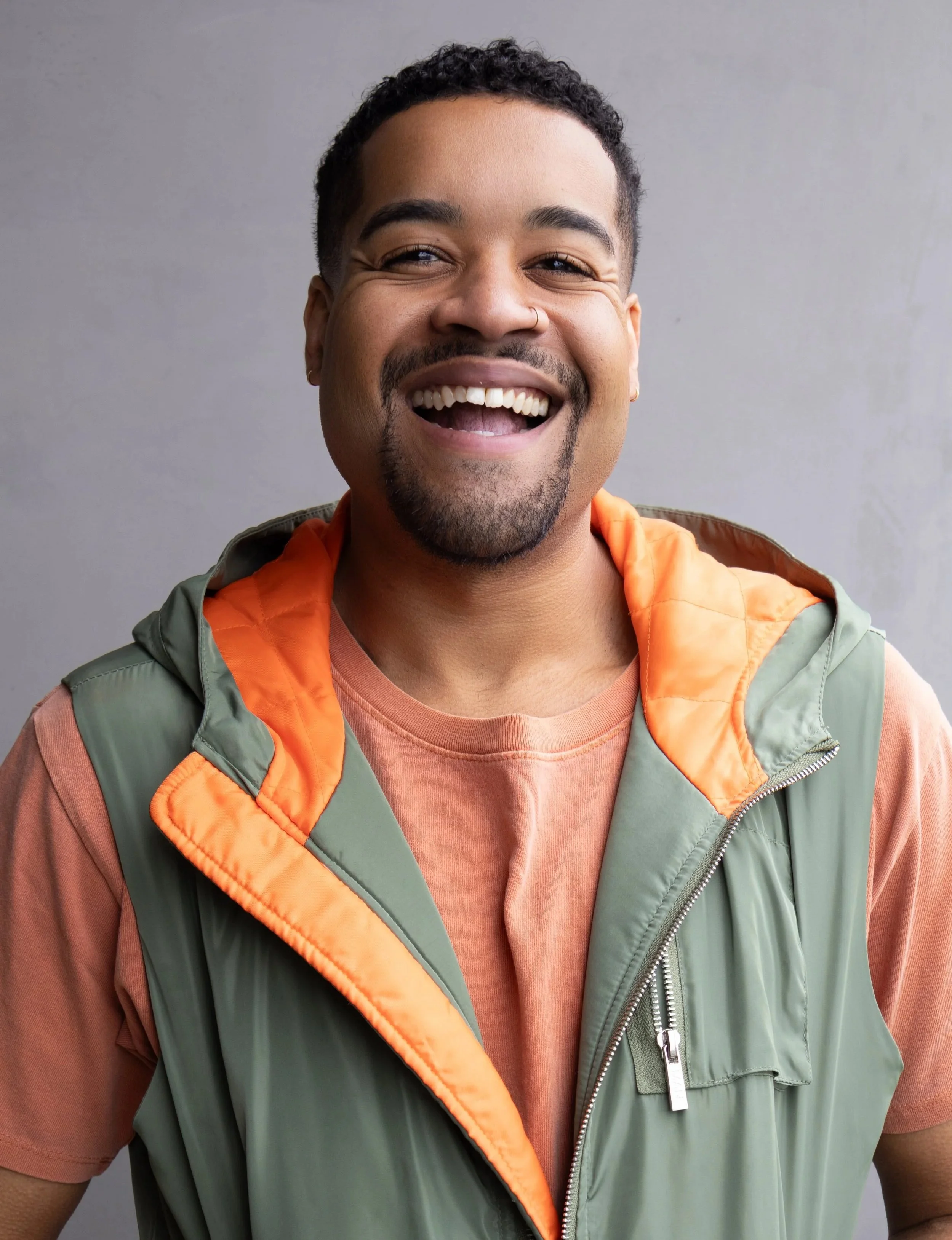 Close-up of a smiling man with short curly hair, a nose ring, and earrings, wearing an orange shirt and a green vest with orange lining, standing against a gray background.