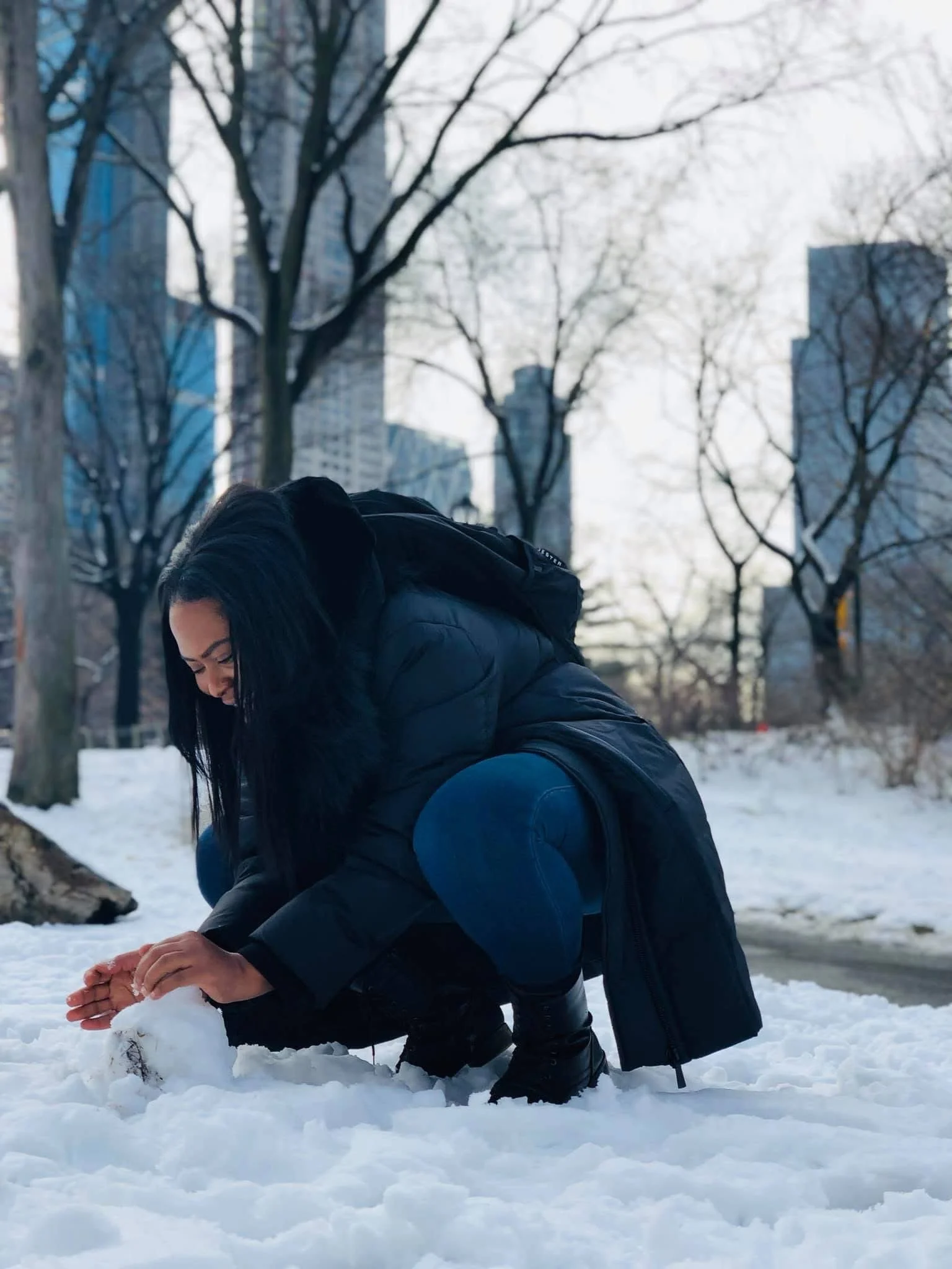 A woman with black hair and glasses is crouching in the snow and building a snowman during winter, with trees and tall buildings in the background.