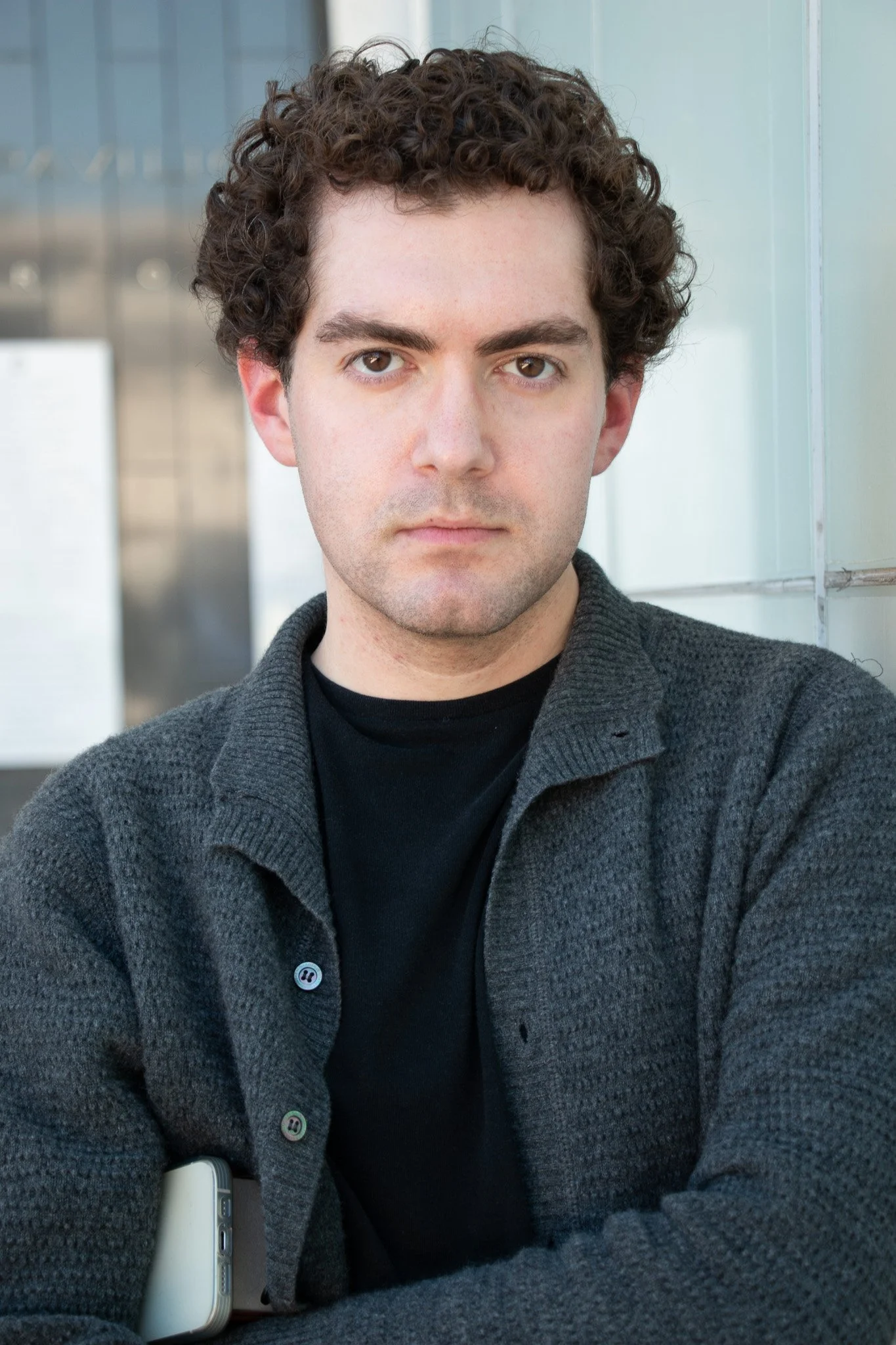 A young man with brown, curly hair and light skin looking directly into the camera. He is wearing a dark shirt and a gray button-up sweater, with a neutral facial expression. The background includes a beige wall with vertical and horizontal lines and an out-of-focus metallic area.