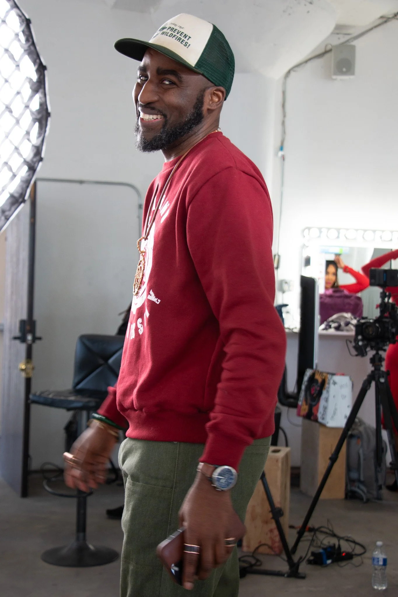 A man wearing a red long-sleeve shirt, green pants, and a trucker hat with green and white mesh, smiling and holding a phone, standing in photography studio with professional lighting, camera, and equipment visible in the background.
