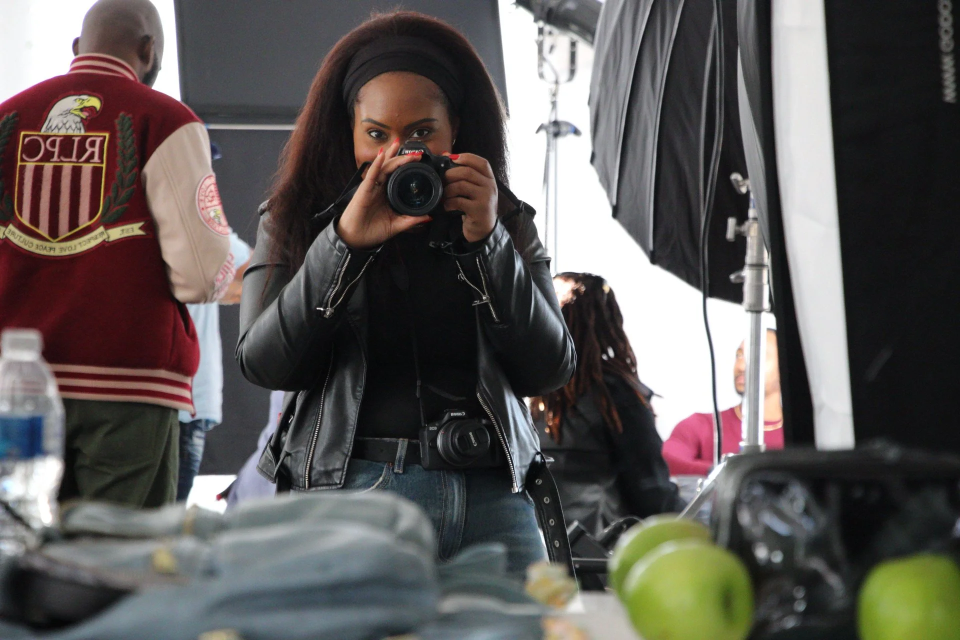 A woman with long dark hair, black headband, and leather jacket taking a photo with a DSLR camera in a busy indoor setting. She has a camera strap around her neck and is looking directly into the camera lens. People and equipment are visible in the background.