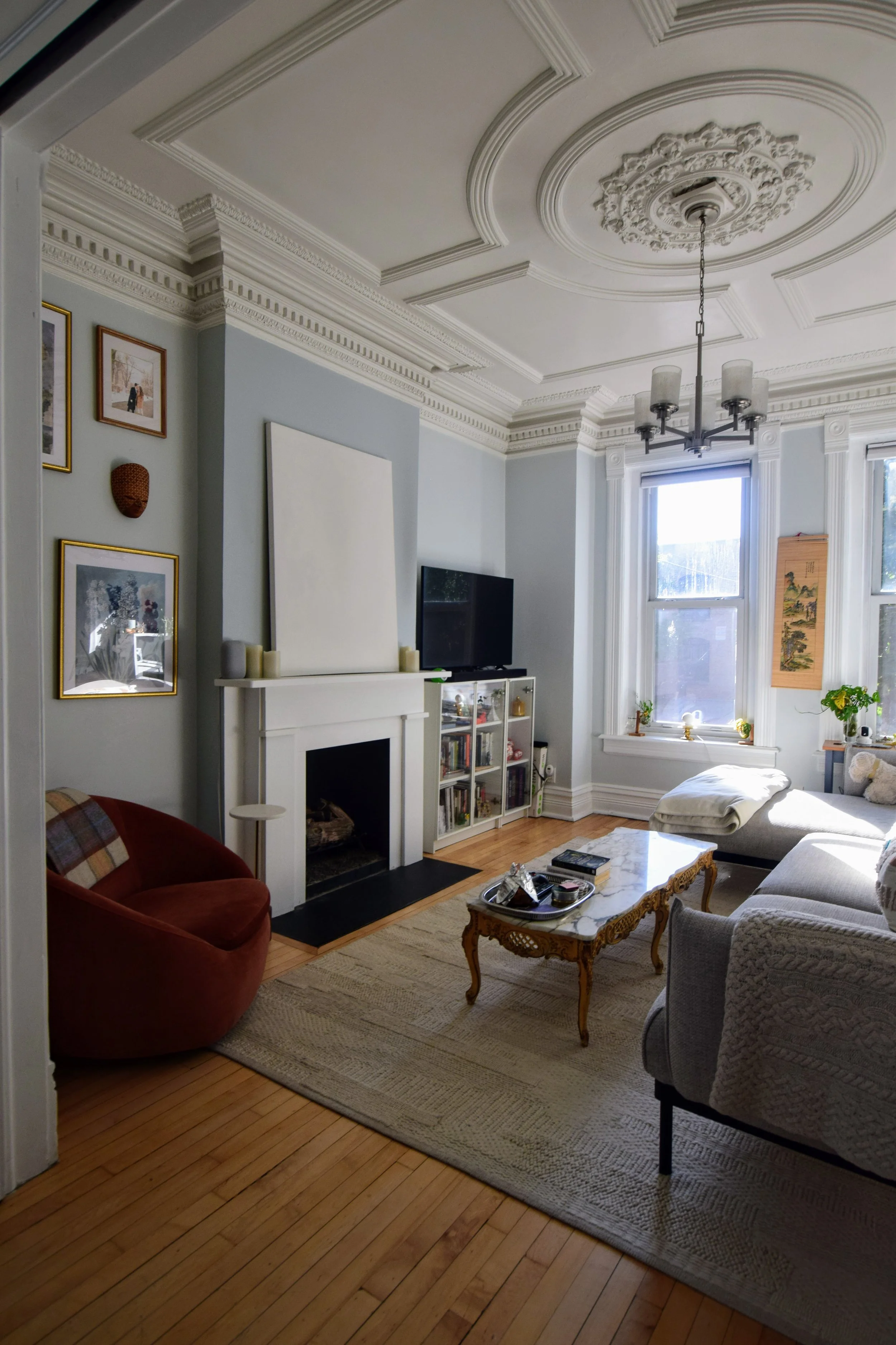 Living room with light blue walls, white ceiling with elaborate molding, and hardwood floors. Contains a red velvet armchair, a beige loveseat, a coffee table with decorative items, a fireplace, a small white cabinet, wall art, and a window with sheer curtains letting in natural light.