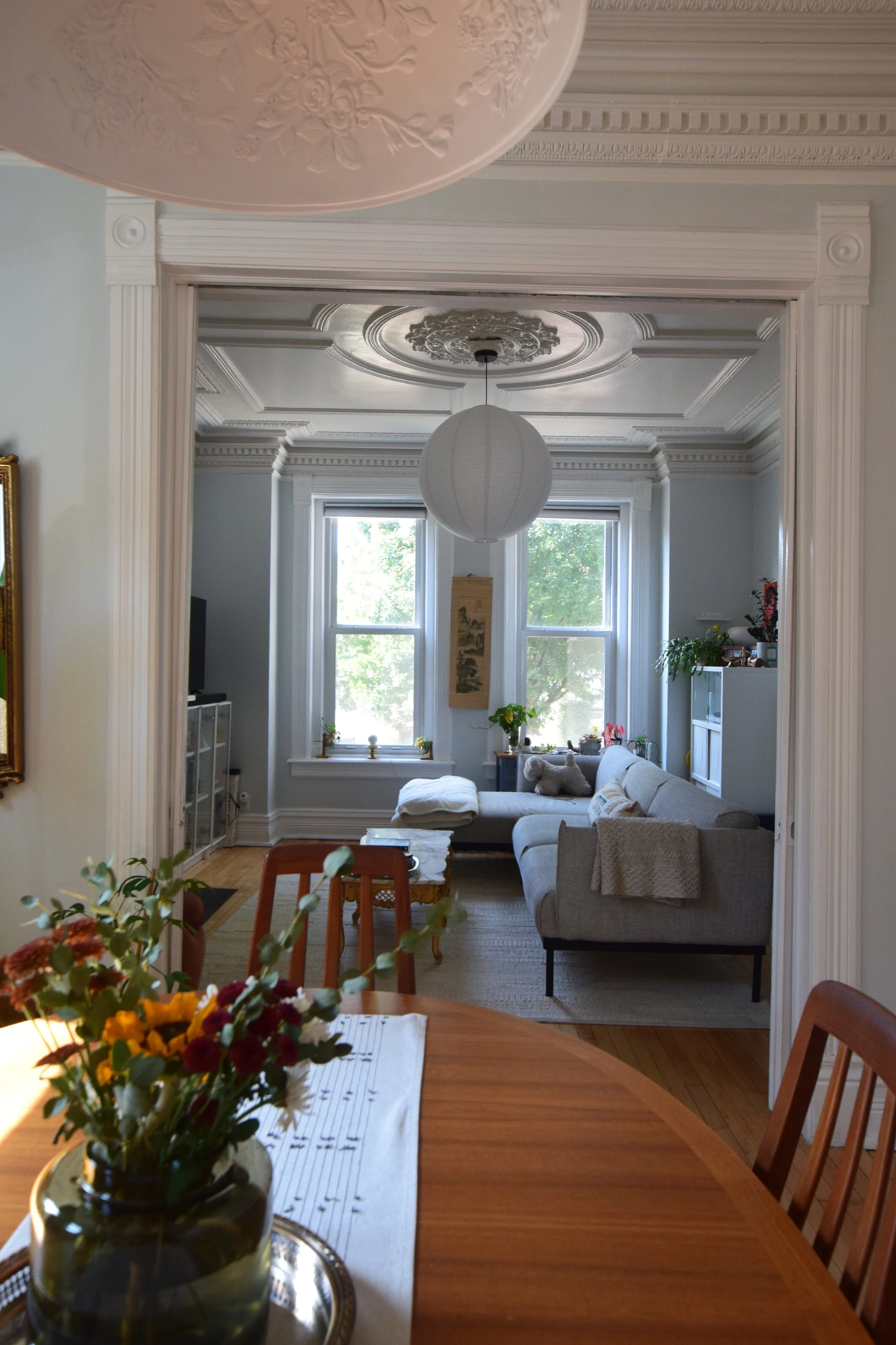 View of a cozy living room with large bay windows, a sofa with cushions, a coffee table, and a white pendant lamp hanging from an ornate ceiling. The room is decorated with plants, artwork, and small decorative items.
