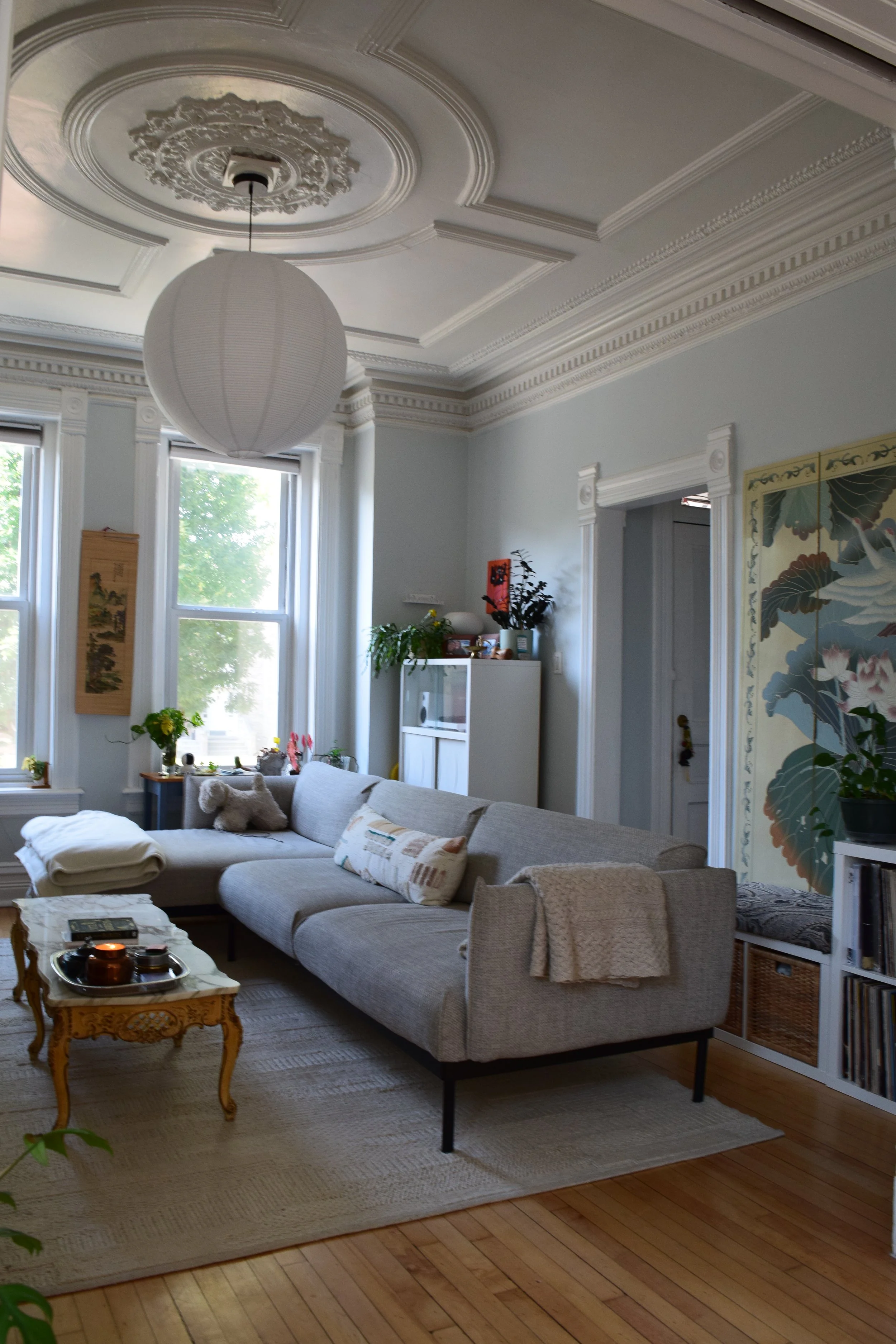 Living room with high ceiling, white walls, large windows, a gray sectional sofa with pillows, a small wooden side table with decorative items, plants, wall art, and traditional molding details.