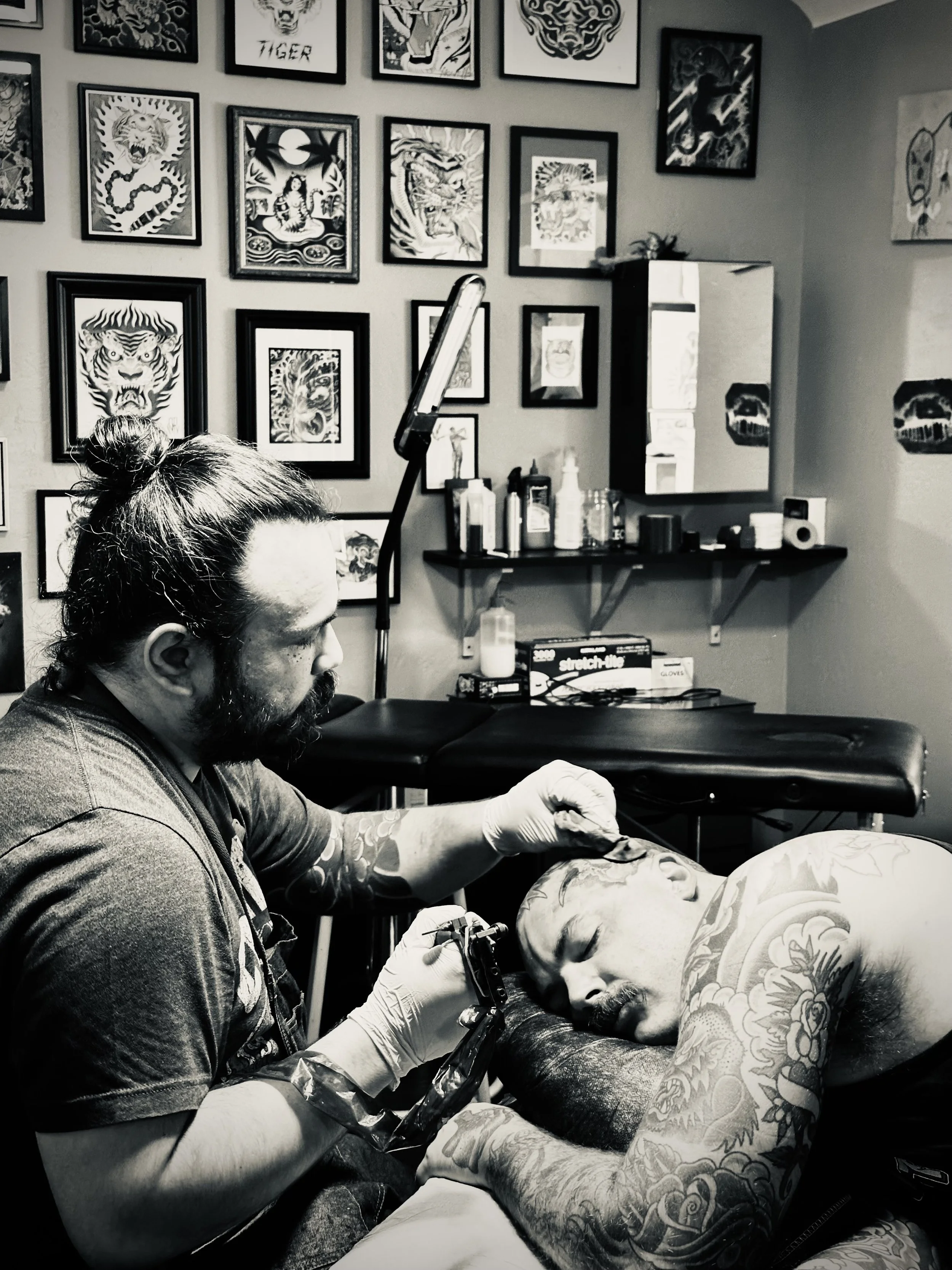 A person with tattoos on their arm and neck receives a tattoo on their arm from a tattoo artist in a tattoo parlor. The parlor has framed tattoo art on the wall and various tattoo supplies on shelves.