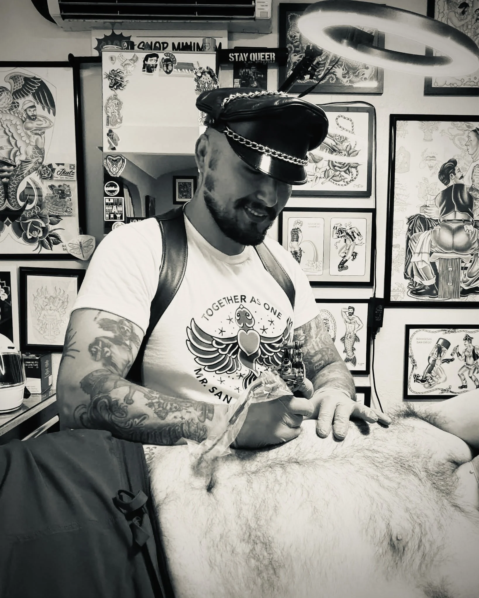 A man with tattoos on his arms, beard, and sunglasses wearing a chain hat accessory, a white t-shirt with a graphic design, and a backpack taking a selfie inside an art gallery with framed tattoos and artwork on the walls.