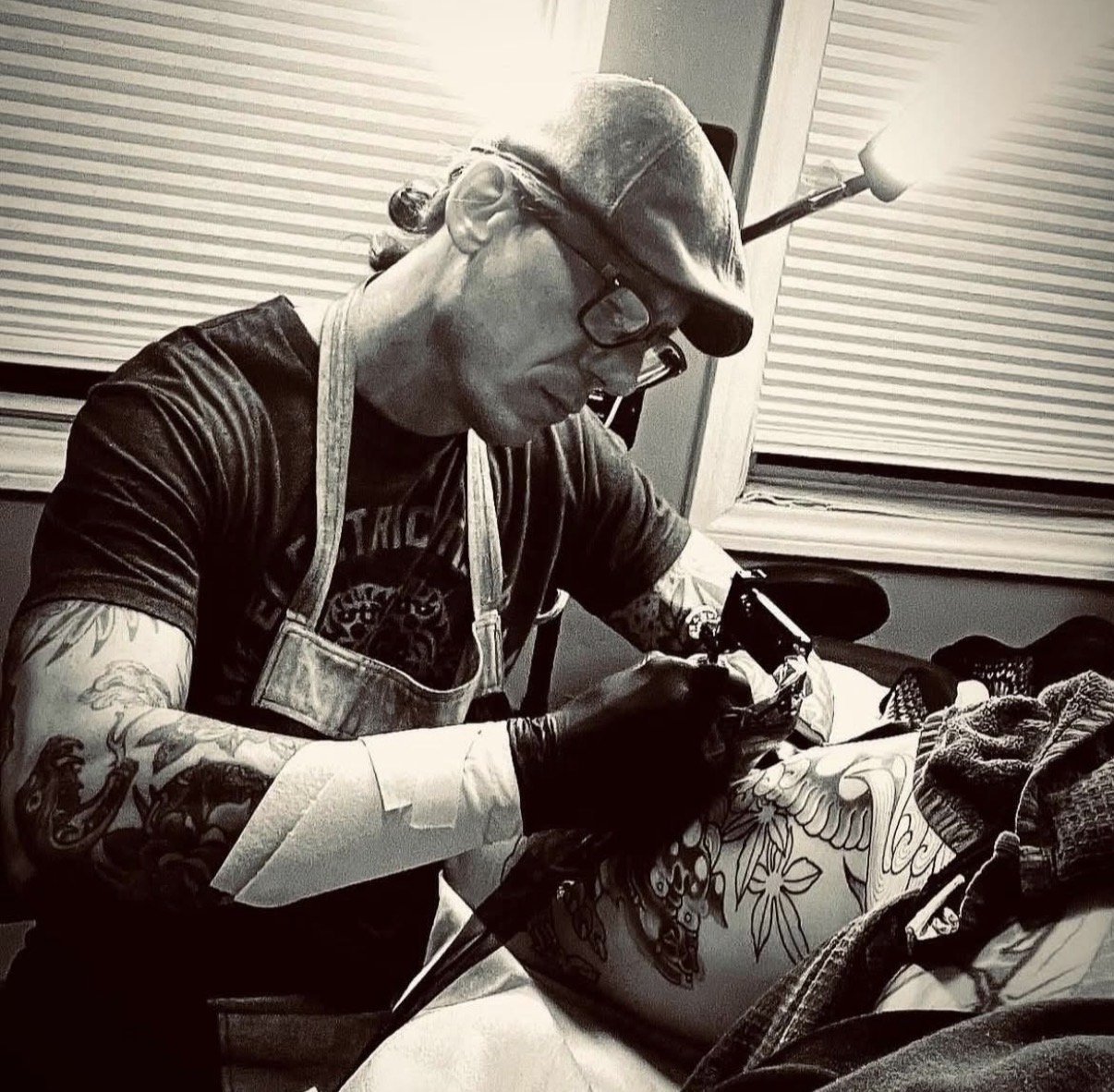 A man wearing glasses, a cap, and a tattooed arm is tattooing someone. The man is wearing gloves and an apron, and is focused on the tattooing process. The room has a window with blinds, and a lamp is providing light.