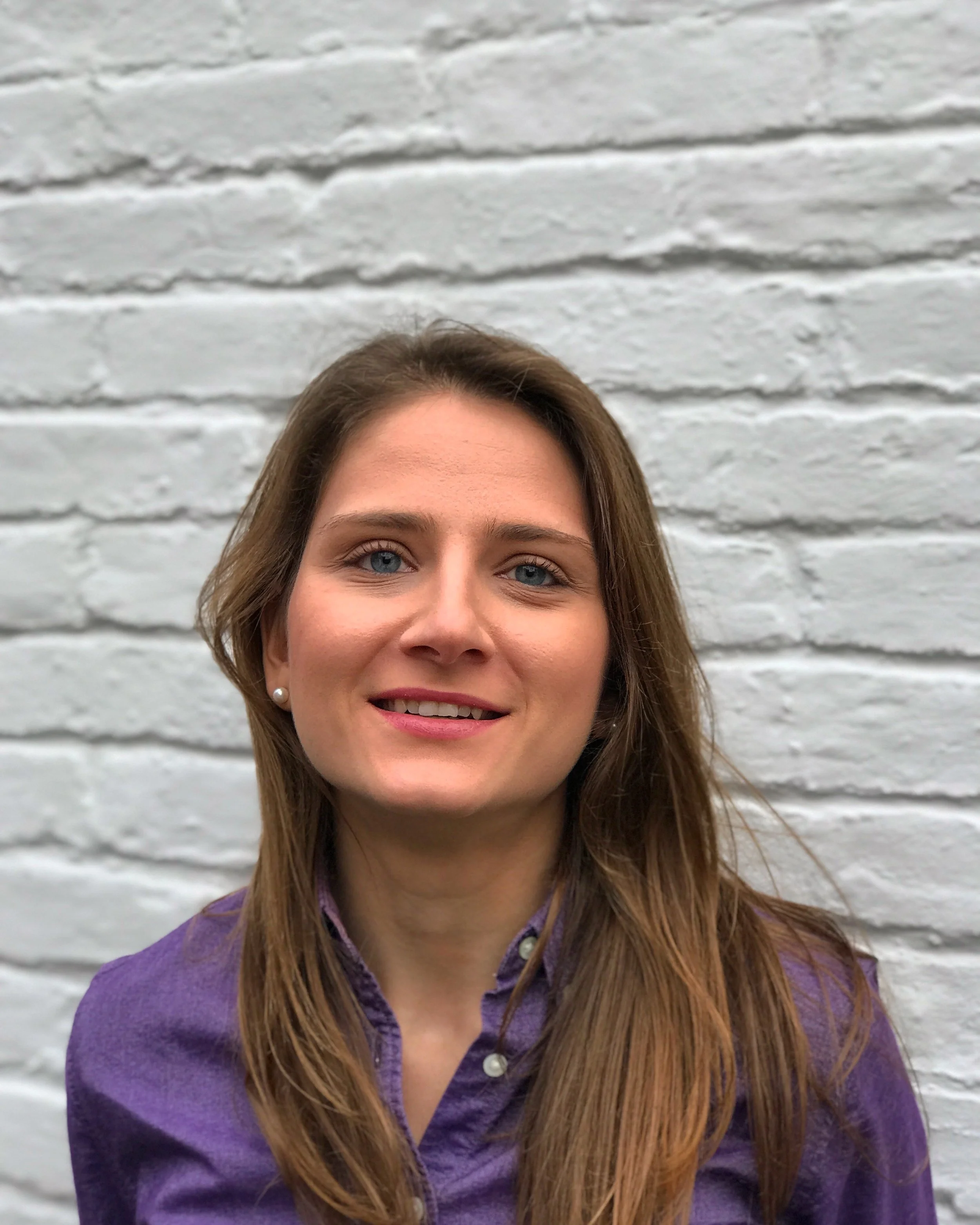 Close-up of a woman with long brown hair, blue eyes, and wearing a purple jacket, standing in front of a white brick wall.