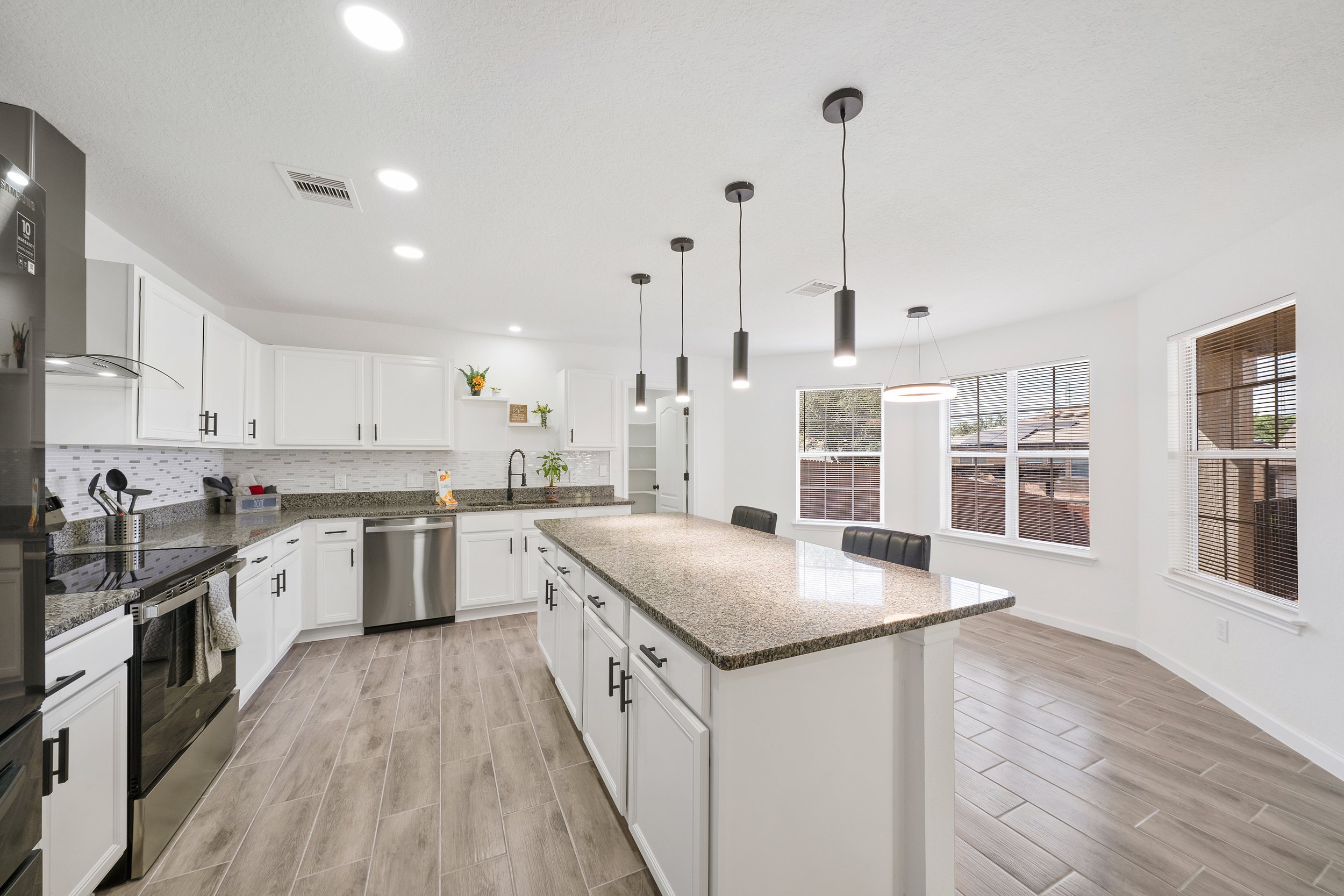 Modern kitchen with white cabinets, granite countertops, hanging pendant lights, large windows with blinds, stainless steel appliances, and wood-look flooring.