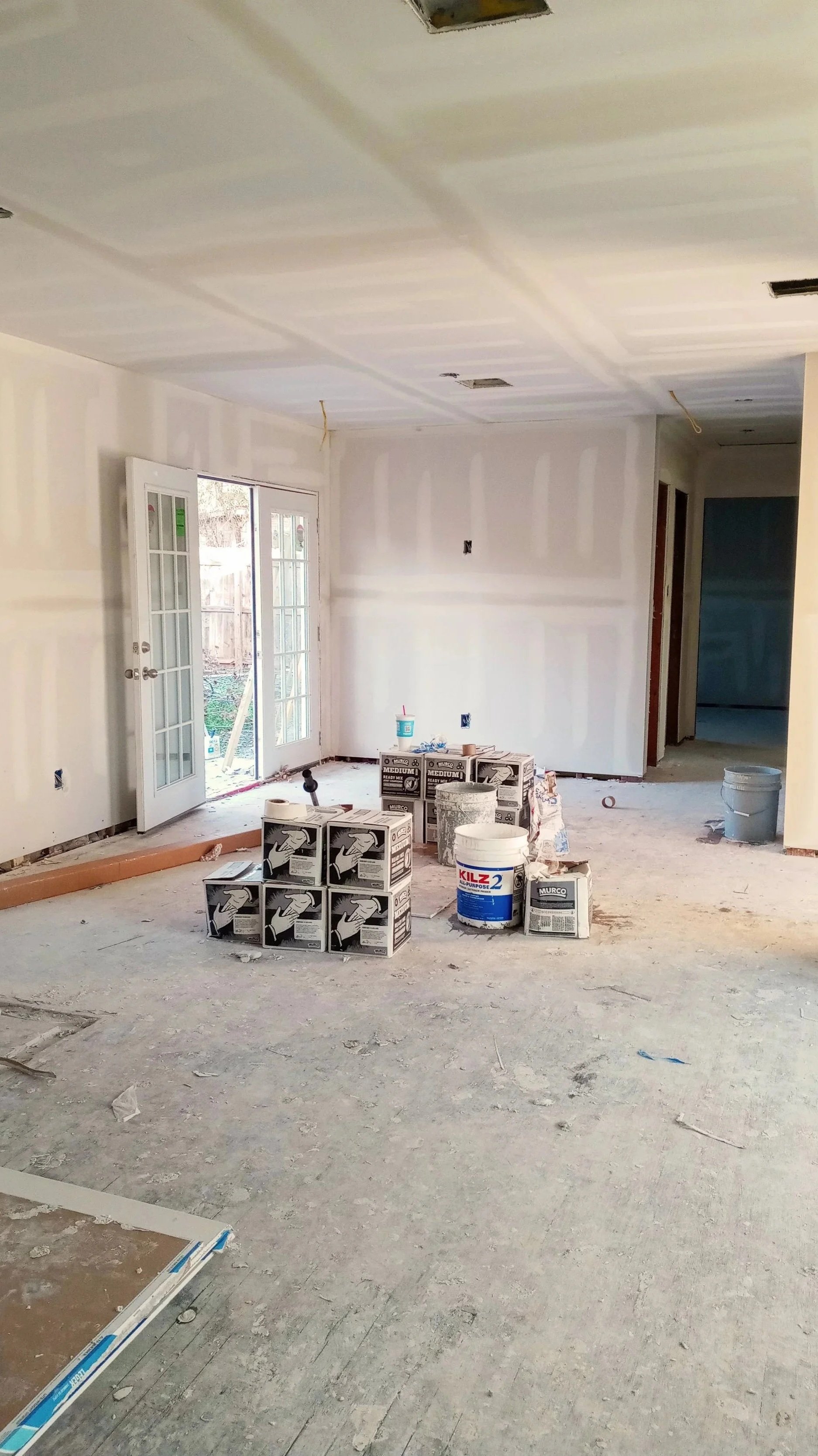 Living room under construction with unfinished walls and ceiling, and various construction materials and supplies on the floor, including boxes of drywall joint compound, buckets, and tools.