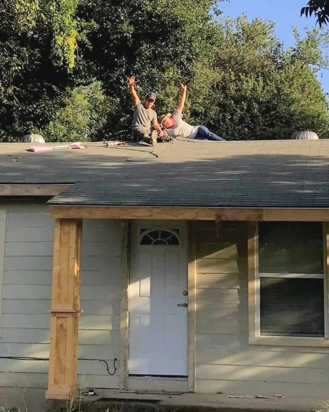Two workers installing or repairing shingles on the roof of a house, with green trees in the background, during daytime.