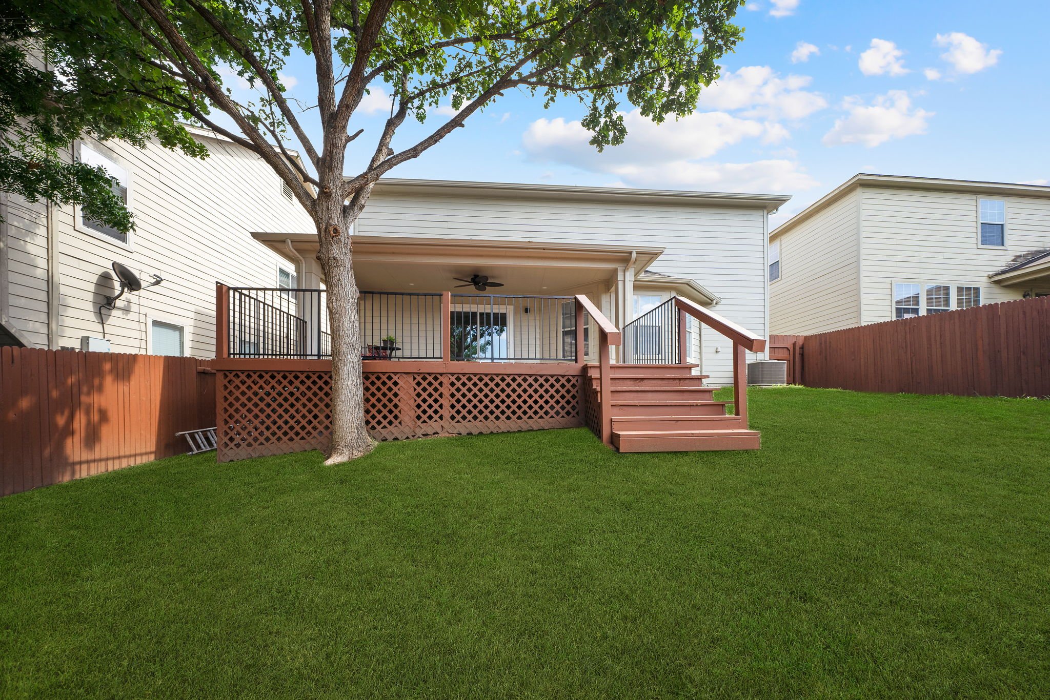 Backyard with a green lawn, a tree, a wooden deck with stairs, and a white two-story house with beige siding, windows, and a door. A wooden fence surrounds the yard.