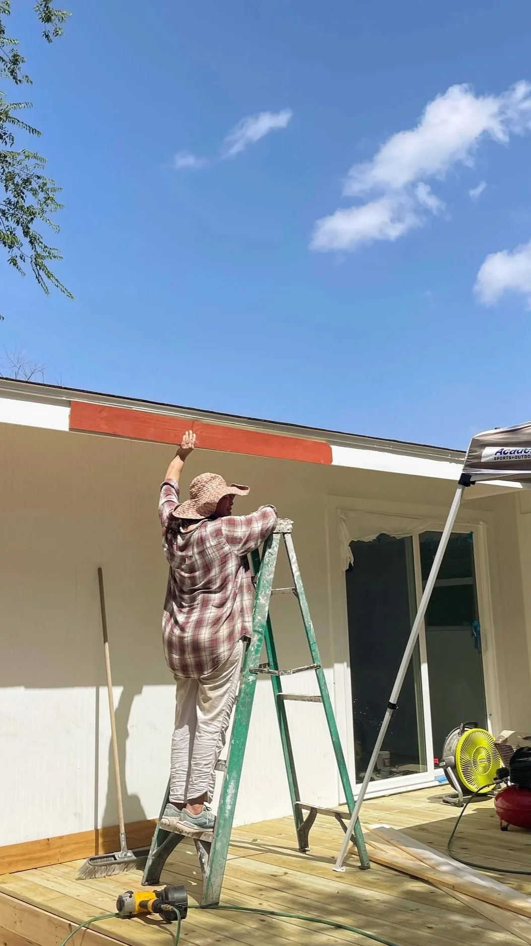 A person standing on a ladder painting the top edge of a house's exterior wall on a sunny day.