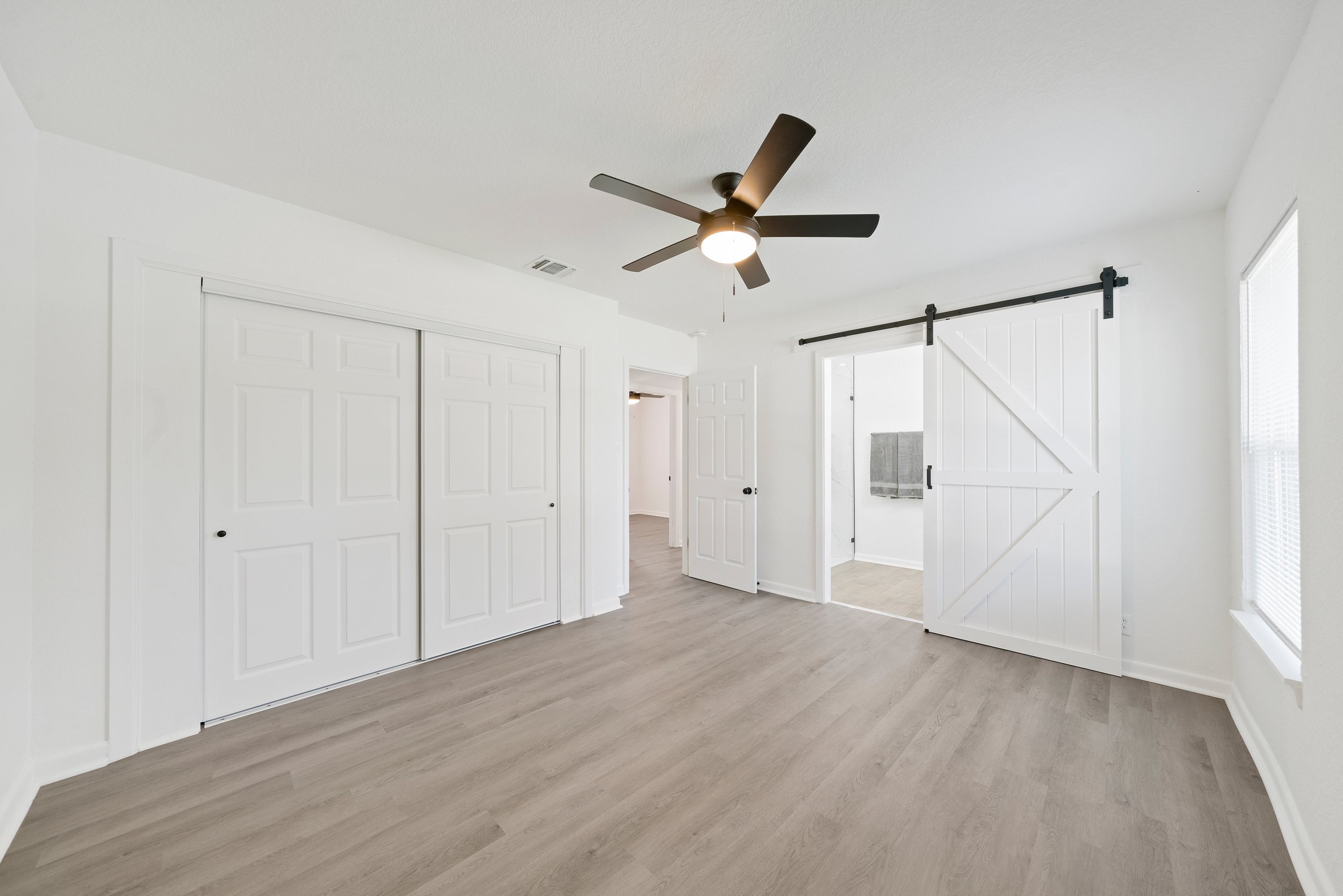 A bright, white room with light-colored wooden flooring, a ceiling fan with a light, a sliding barn door, closed closet doors, and large windows with blinds.