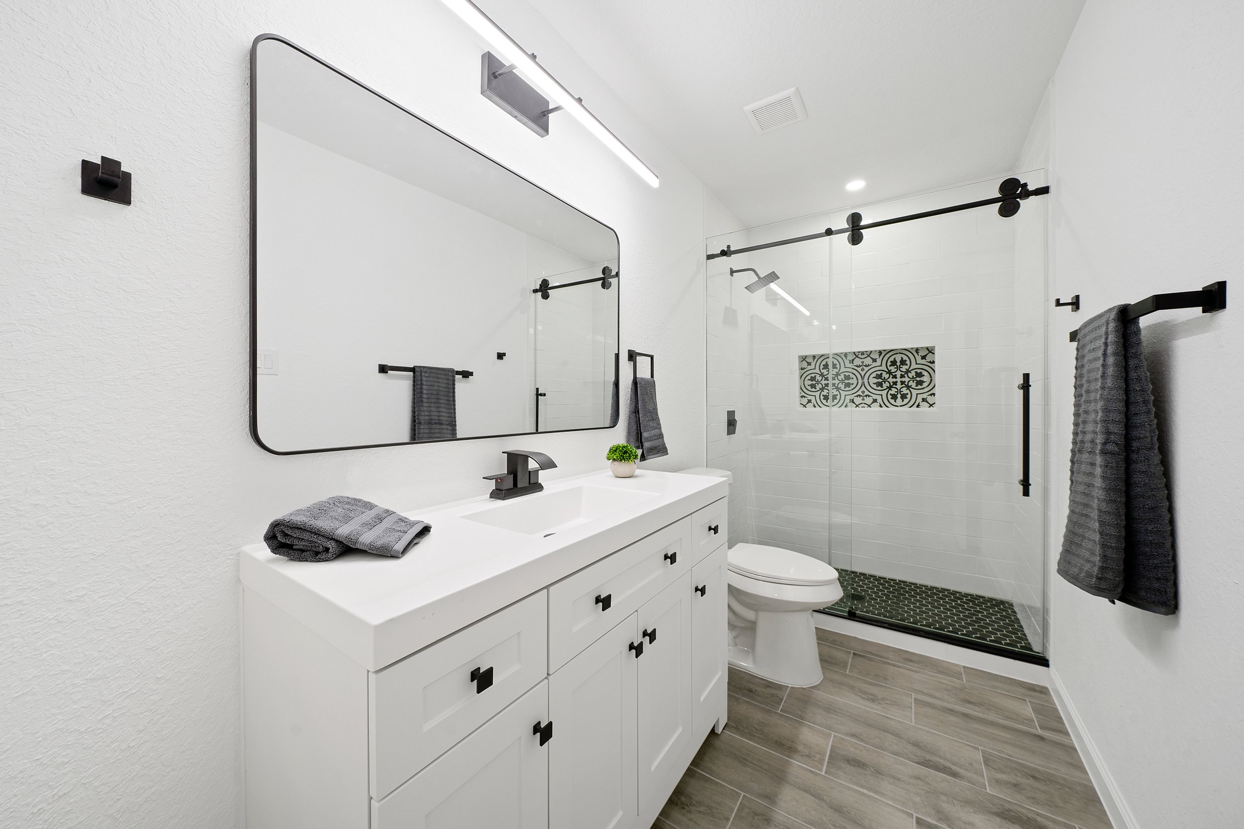 Modern bathroom with white vanity, black fixtures, large mirror, gray towels, plant, shower with glass door, patterned tile accent, and wood-look floor.