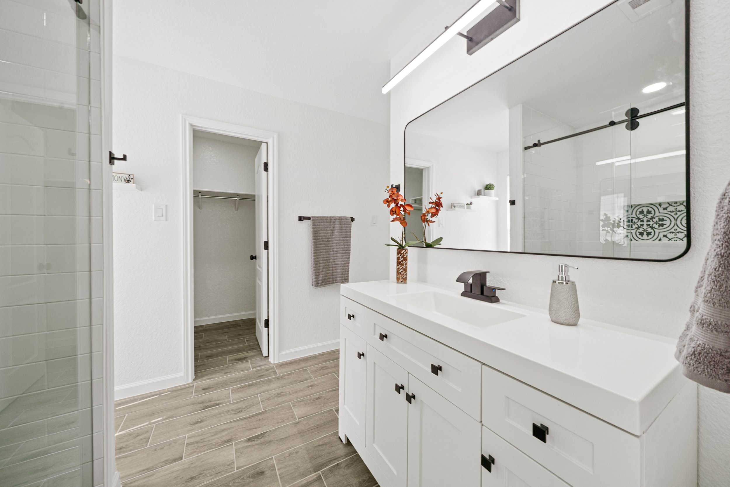 Modern bathroom with white vanity, large mirror, flower vase, faucet, and towels, with a closet door open and decorative tiles in shower area.