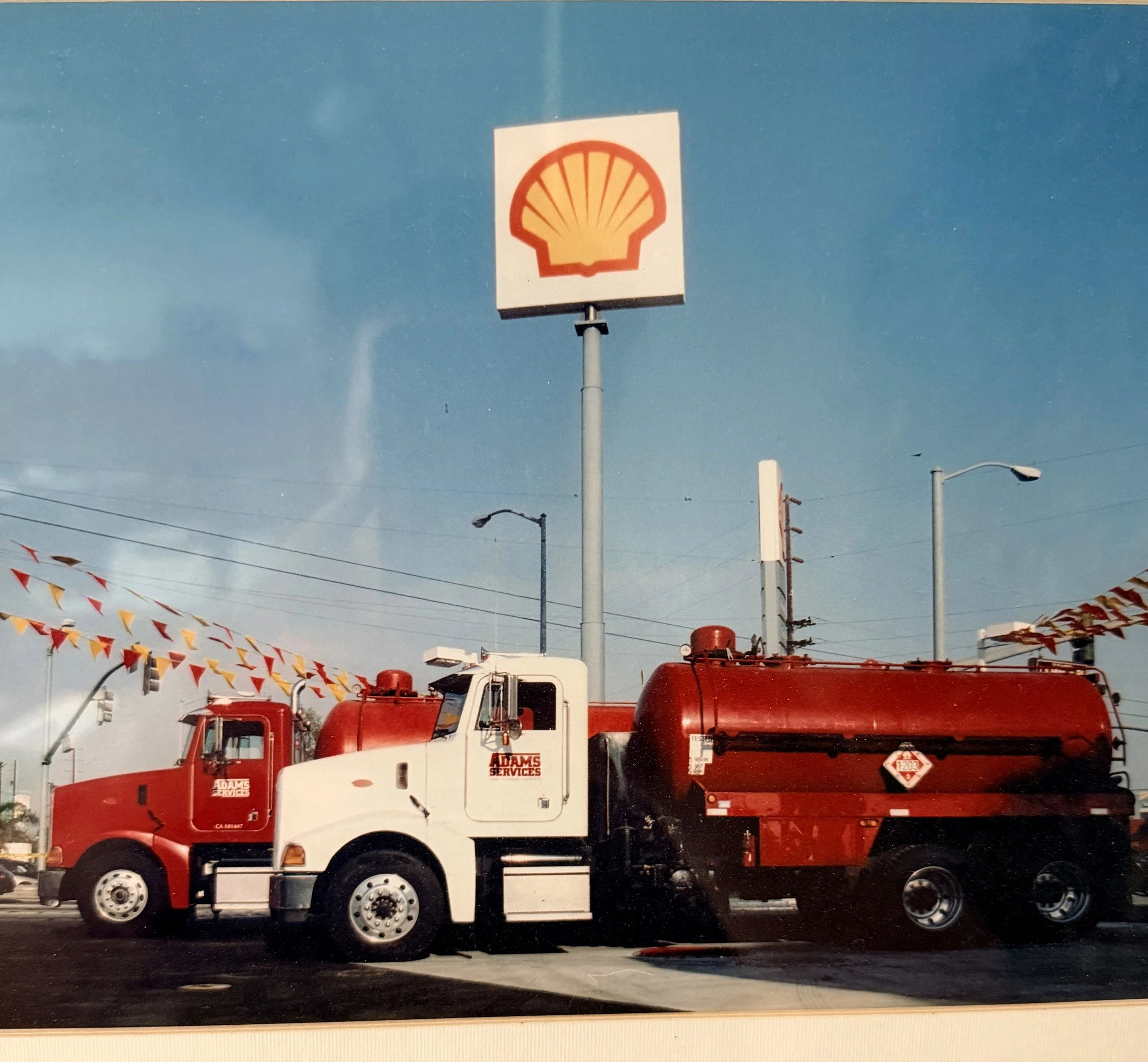 A row of large red and white utility trucks parked outdoors, with a sign displaying the Shell logo and various traffic flags hanging overhead against a blue sky.